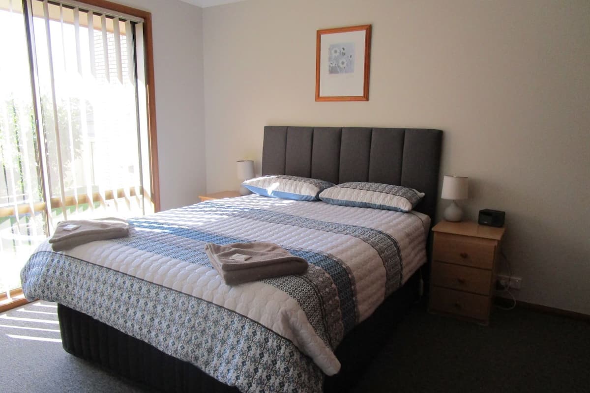 A neatly made bed with a patterned bedspread, a bedside table with a lamp, and a framed picture on the wall.
