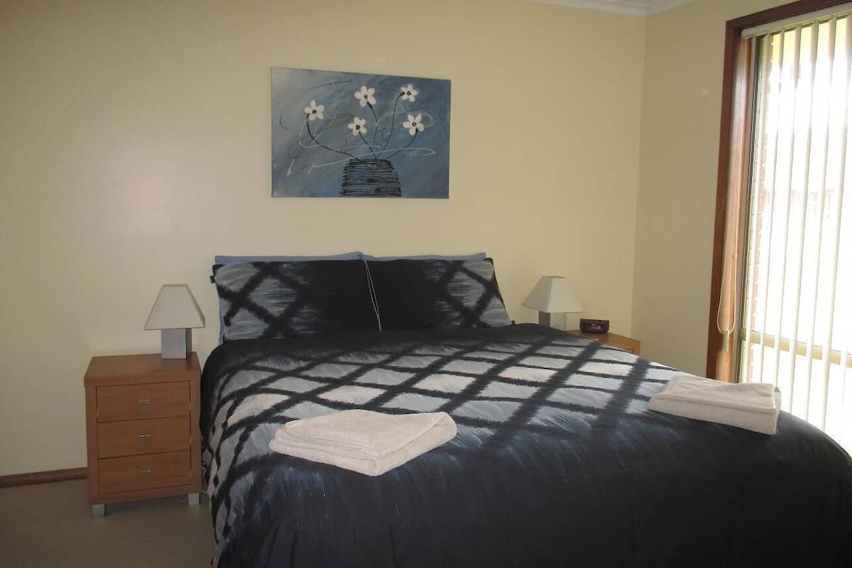 A bedroom with a bed, a nightstand, a lamp, a painting, and a window with blinds.