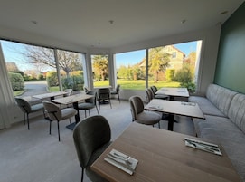 Un espace de repas moderne, doté de tables et de chaises, d’un canapé et de grandes fenêtres offrant une vue sur un jardin et des bâtiments.