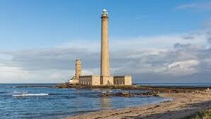 Um farol com uma torre alta e uma construção mais baixa nas proximidades, localizado numa costa rochosa com o mar ao fundo.