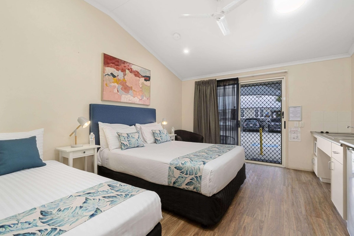 A hotel room with two beds, a desk, a chair, a ceiling fan, and a view of the street through a sliding door.