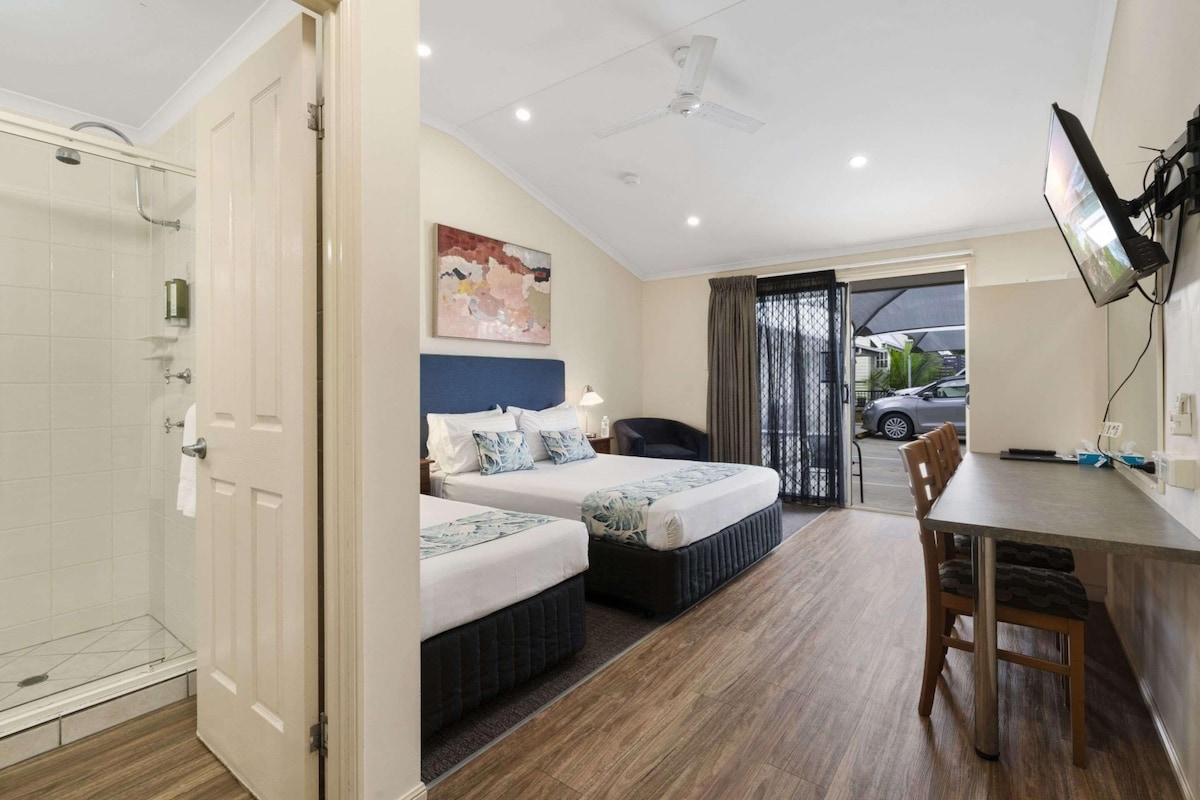 A hotel room with two beds, a desk, a chair, a TV, a shower, and a view of the outside.
