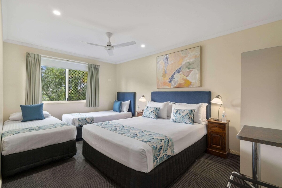 A hotel room with three beds, a ceiling fan, a large window with curtains, a painting on the wall, and a small table with a lamp.