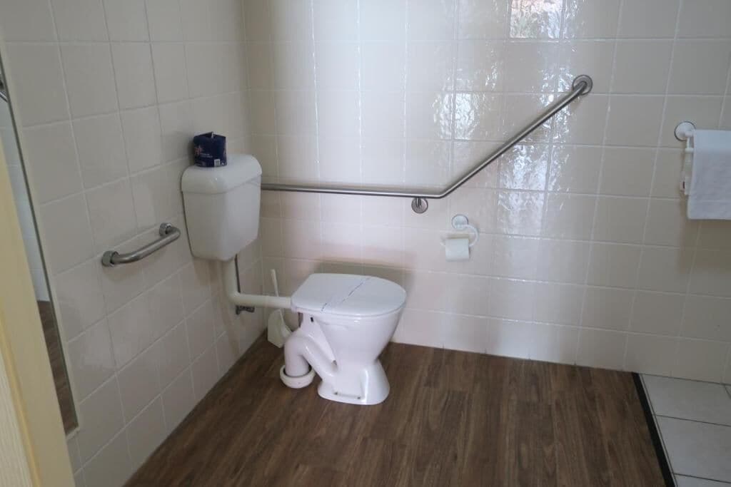 Un baño con inodoro blanco, barra de apoyo y pared con azulejos blancos.