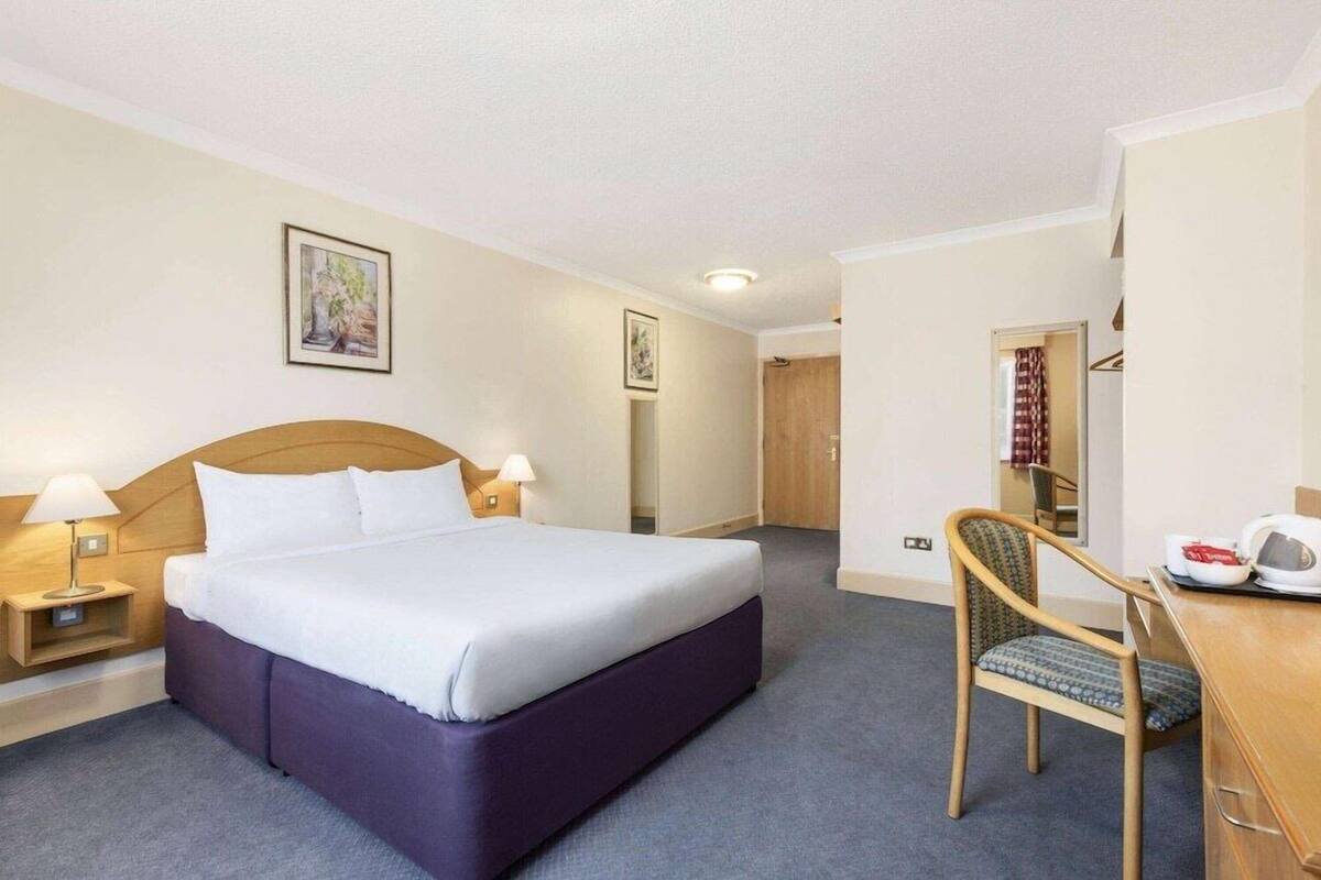 Ett hotellrum med en säng, sänglampor, ett skrivbord med en stol och en dörr som leder till ett annat rum.