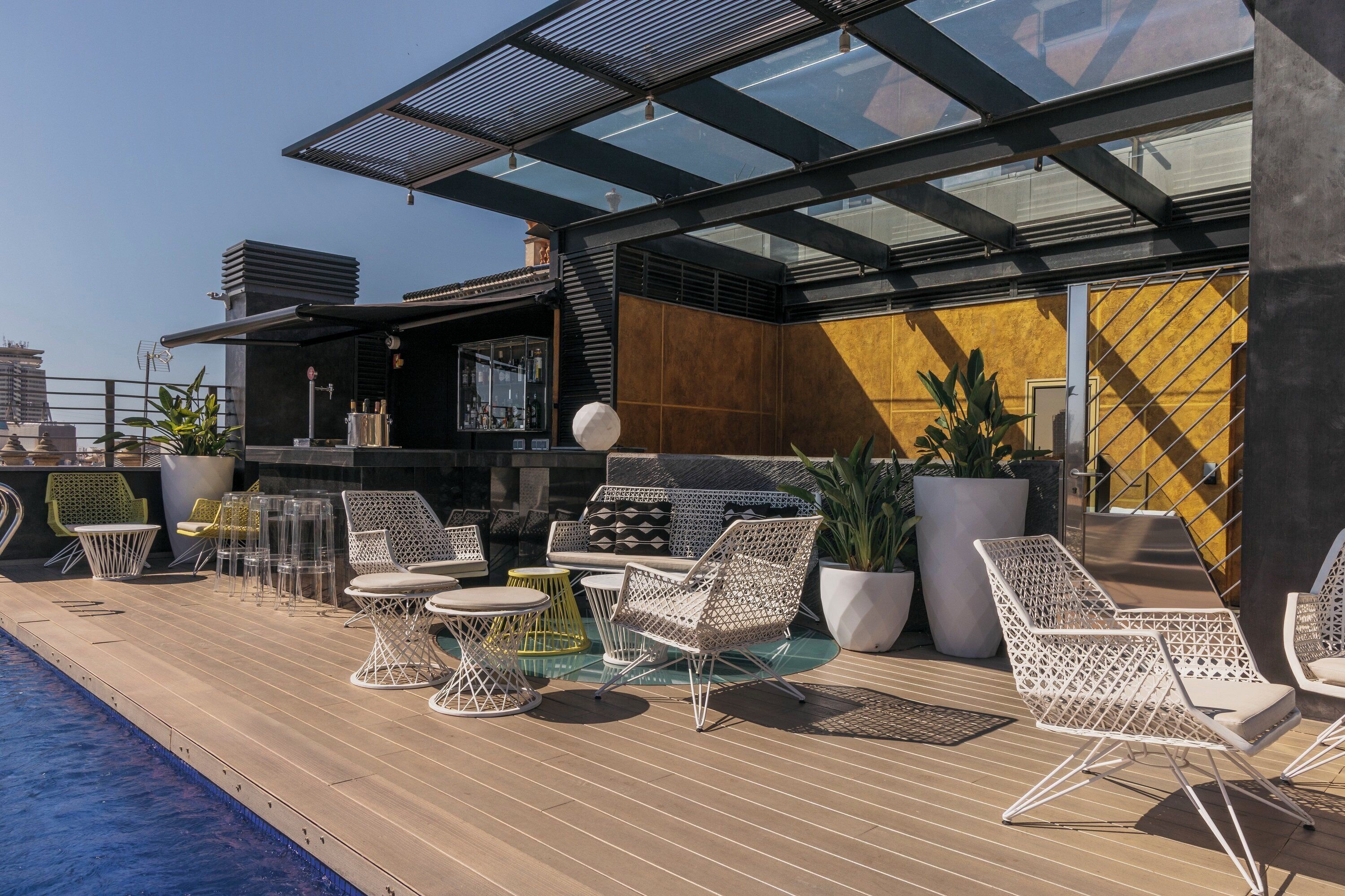 A rooftop bar with outdoor seating, including white wicker chairs and a glass table, overlooking a cityscape.