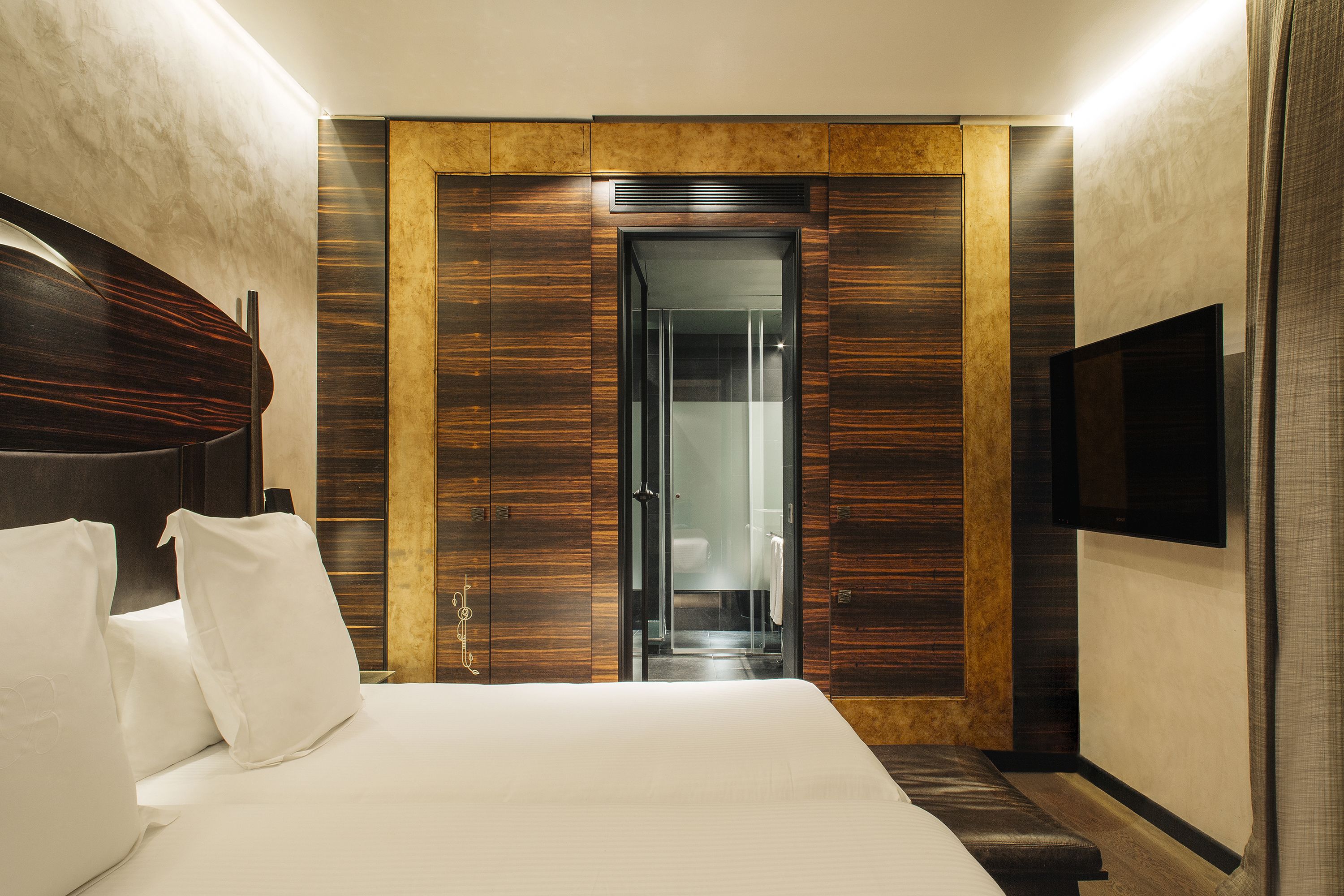 Una habitació d'hotel moderna amb un llit gran, un armari de fusta, una porta de vidre que condueix a un bany i un televisor penjat.