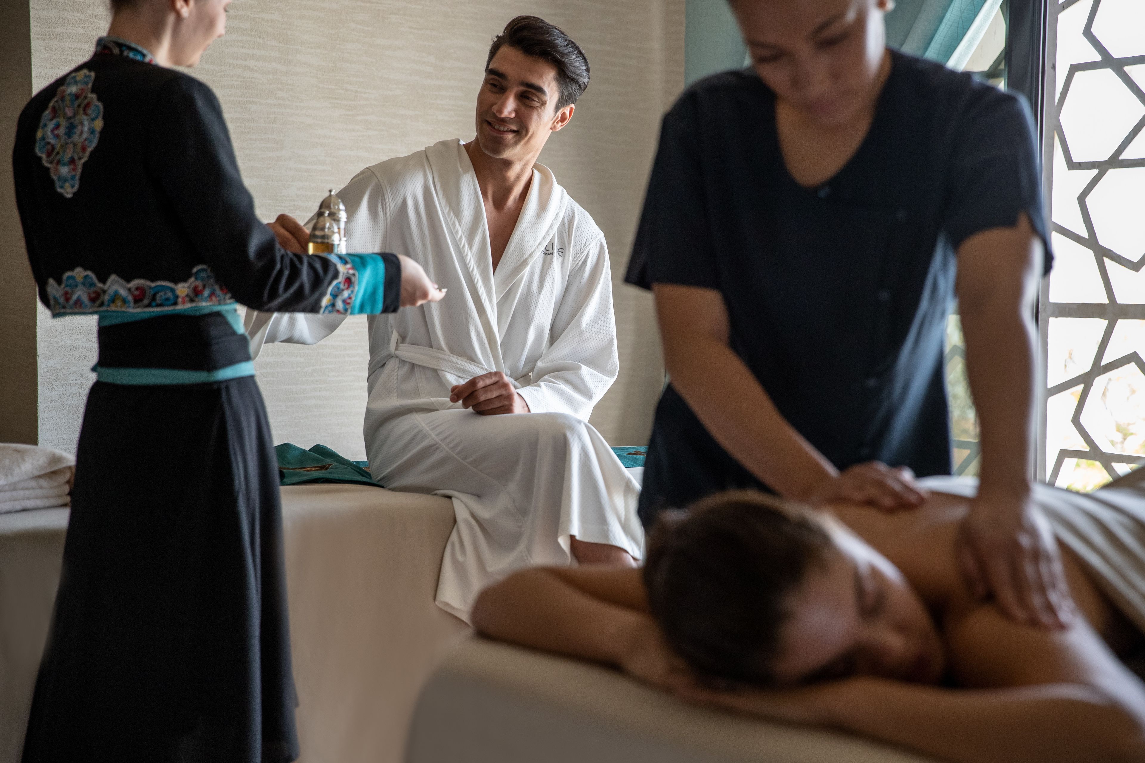 A massage therapy session in a room with a bed, a massage table, and a window with a geometric pattern.