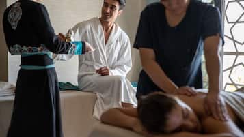 A massage therapy session in a room with a bed, a massage table, and a window with a geometric pattern.