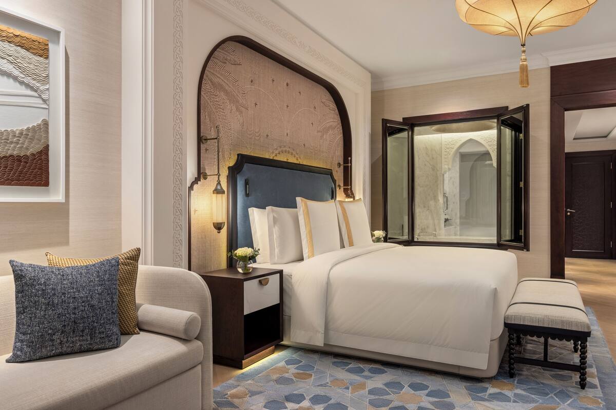 Une chambre avec un grand lit, une table de chevet, une chaise et une grande fenêtre.