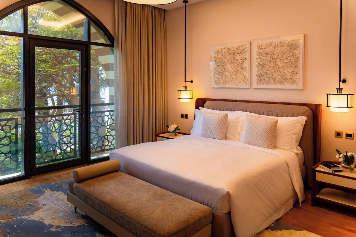Une chambre d’hôtel avec un grand lit, deux tables de chevet, un banc, une grande fenêtre donnant sur un beau paysage et deux lampes fixées au mur.