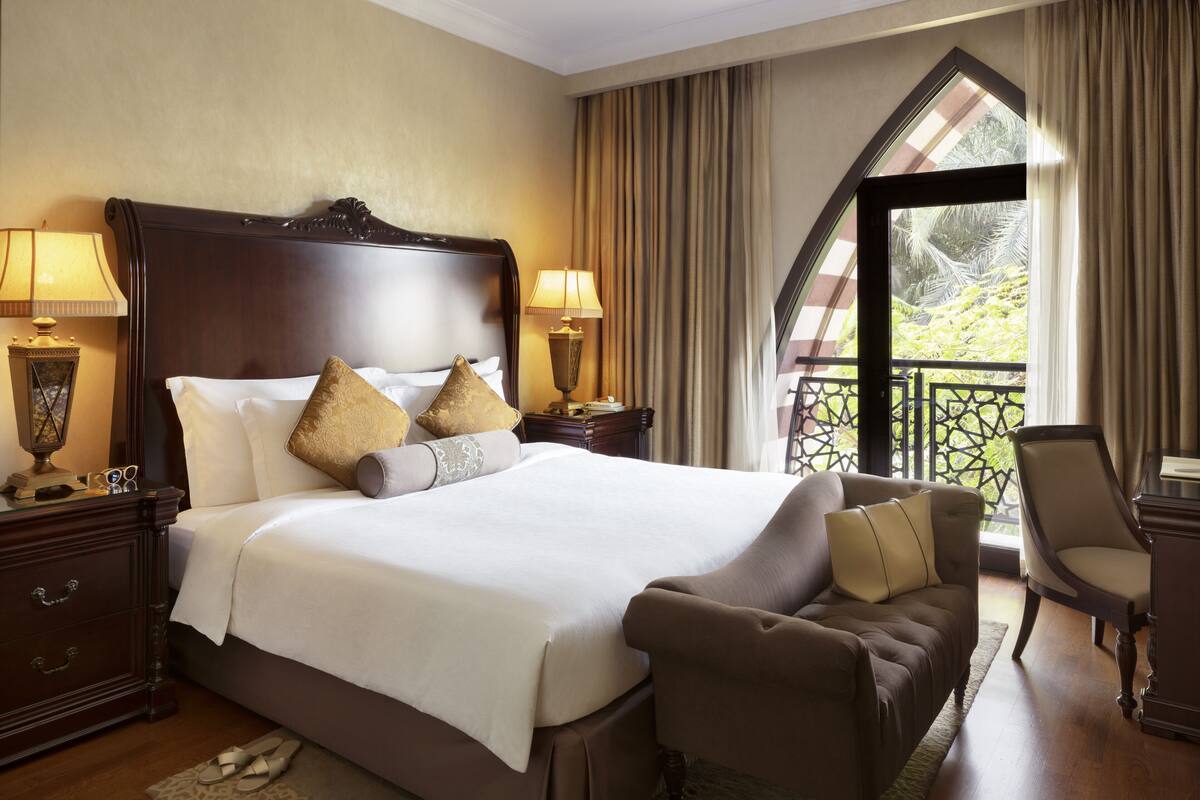 Une chambre d’hôtel avec un grand lit, deux tables de chevet avec des lampes, un canapé et une vue sur la verdure par la fenêtre.