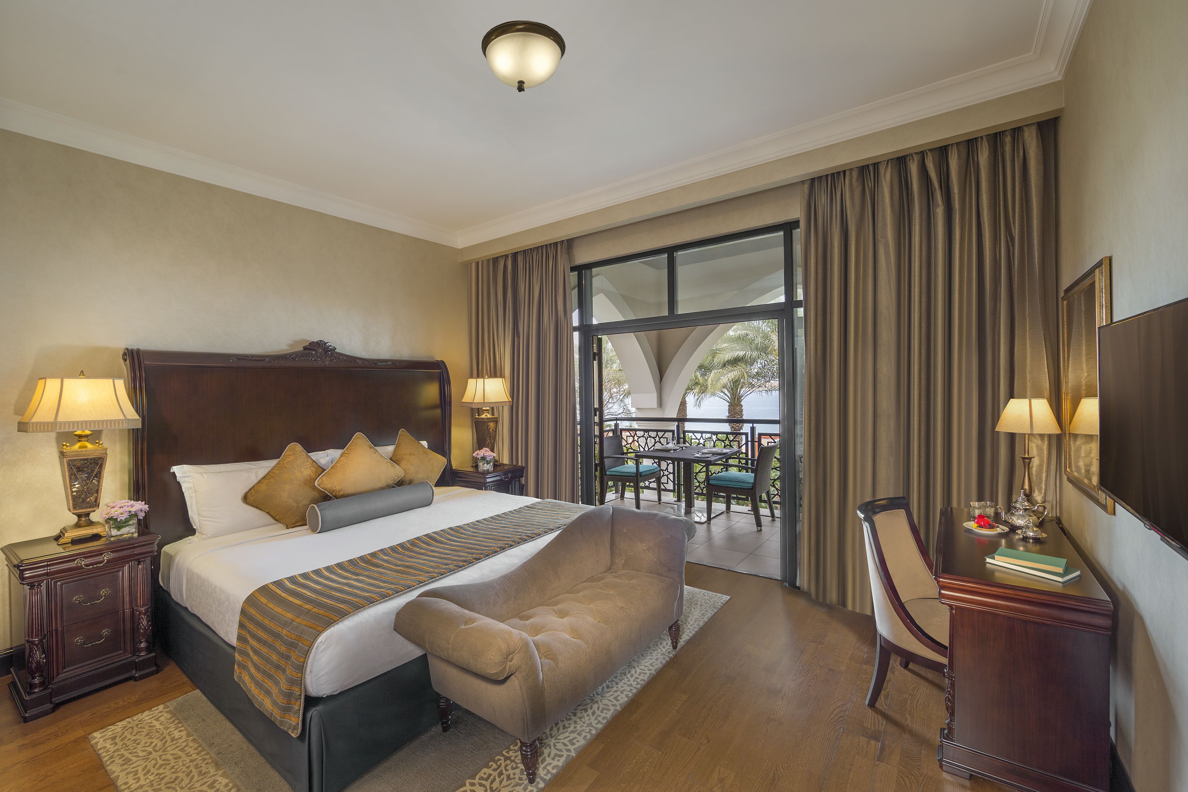 A hotel room with a large bed, two bedside tables with lamps, a chair, a desk, and a balcony with a table and chairs.