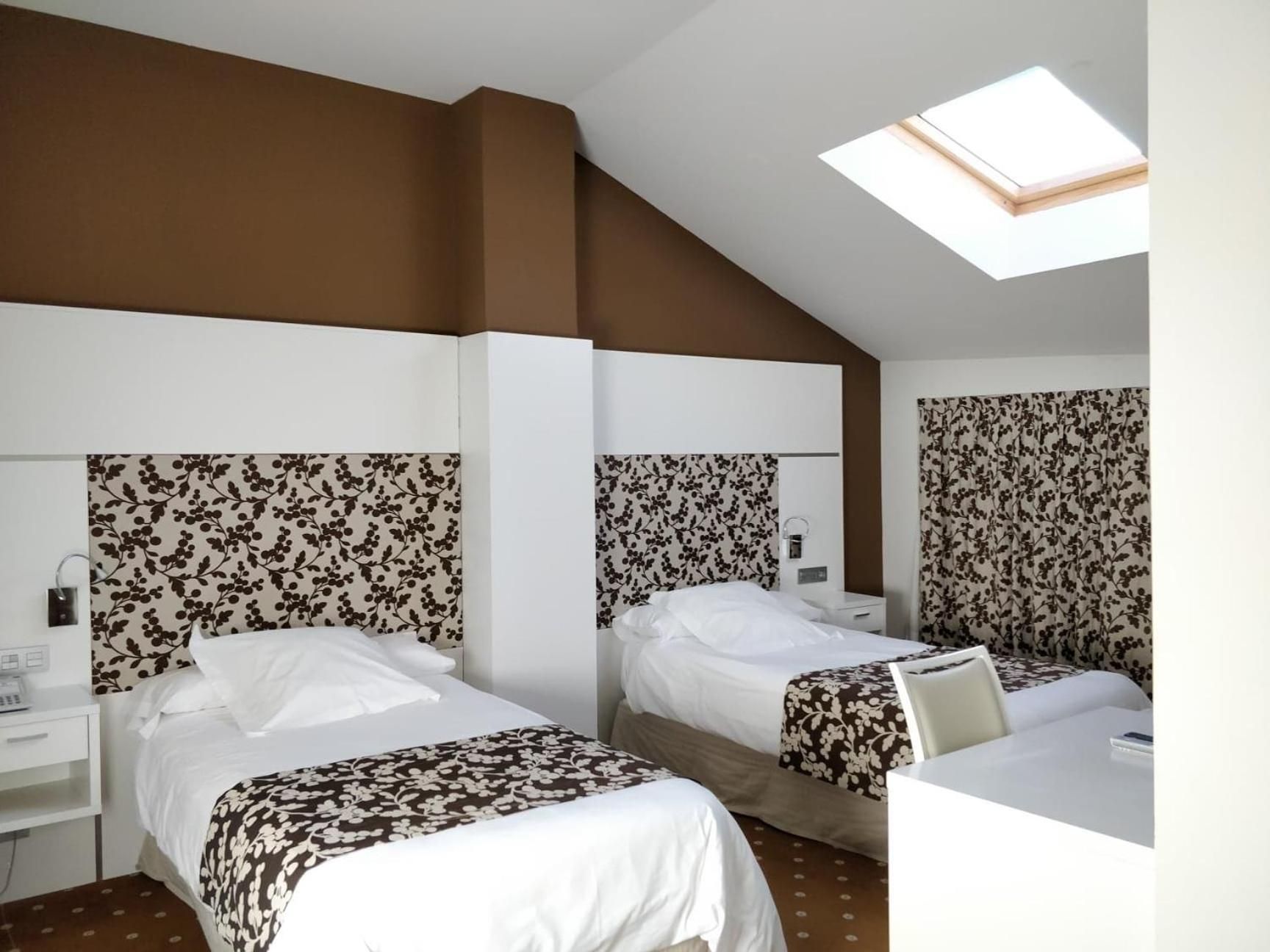 A hotel room with two beds, a desk, a chair, and a skylight.