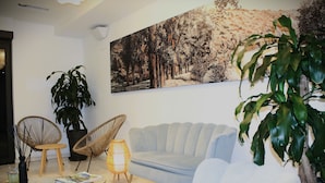 Uma sala de estar com um sofá azul claro, duas cadeiras de vime, uma mesa de centro de vidro, uma grande planta em vaso e um quadro pendurado na parede.