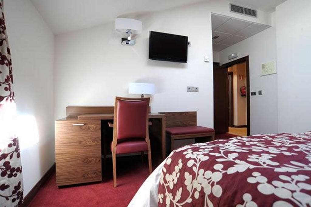 A hotel room with a bed, a desk with a chair, a television mounted on the wall, and a door leading to another room.