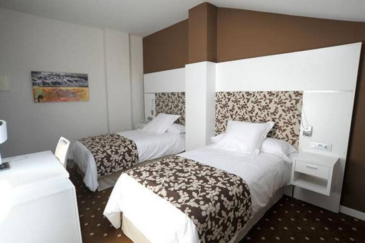 Habitación de hotel con dos camas, cada una con cabecero estampado con motivos florales, ropa de cama blanca y una mesita de noche pequeña con lámpara.
