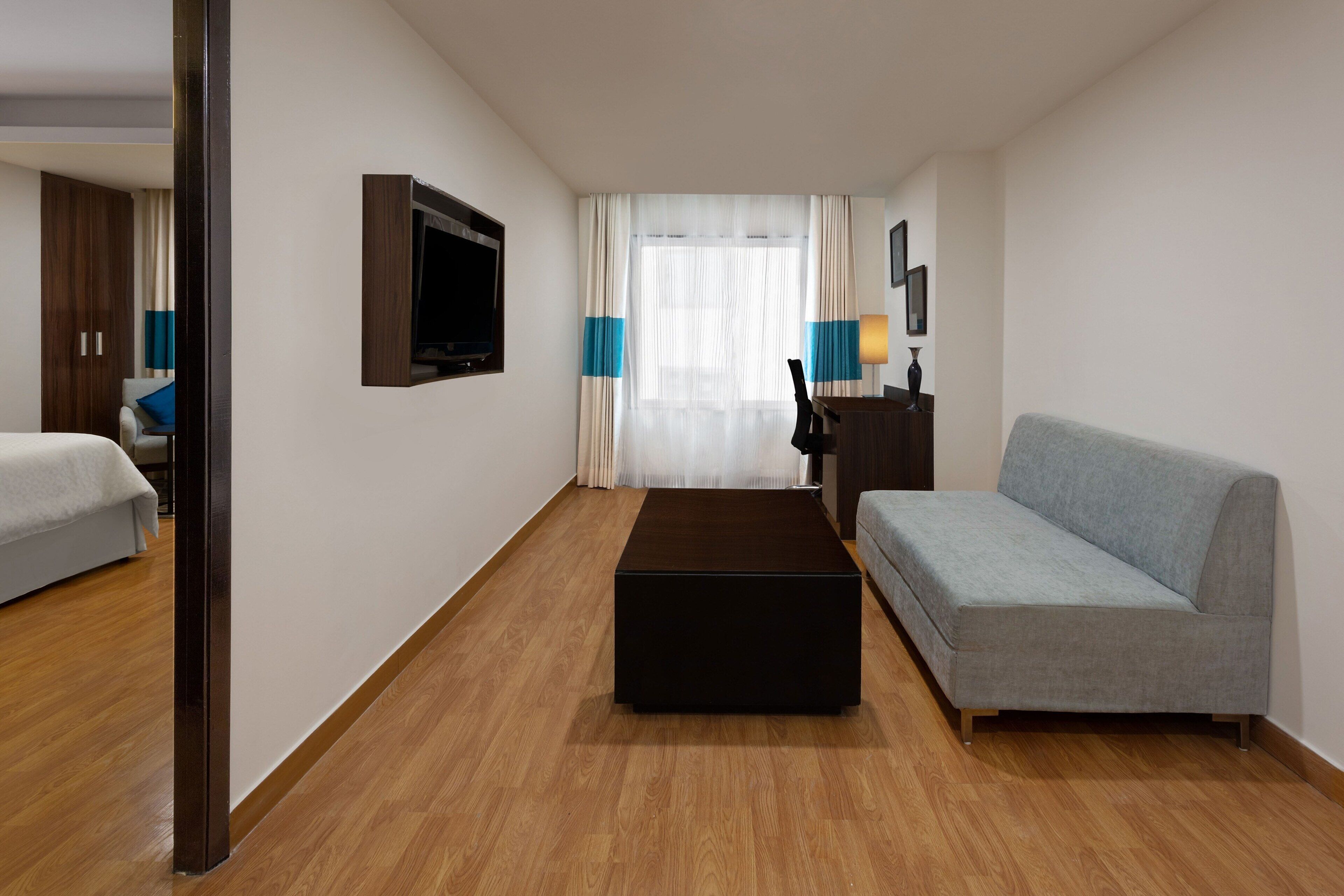 A modern hotel room with a wooden floor, a grey sofa, a black ottoman, a TV mounted on the wall, and a large window with sheer curtains.