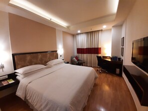A hotel room with a large bed, a desk, a chair, a TV, and a window with curtains. - Four Points by Sheraton Vadodara (Vadodara)