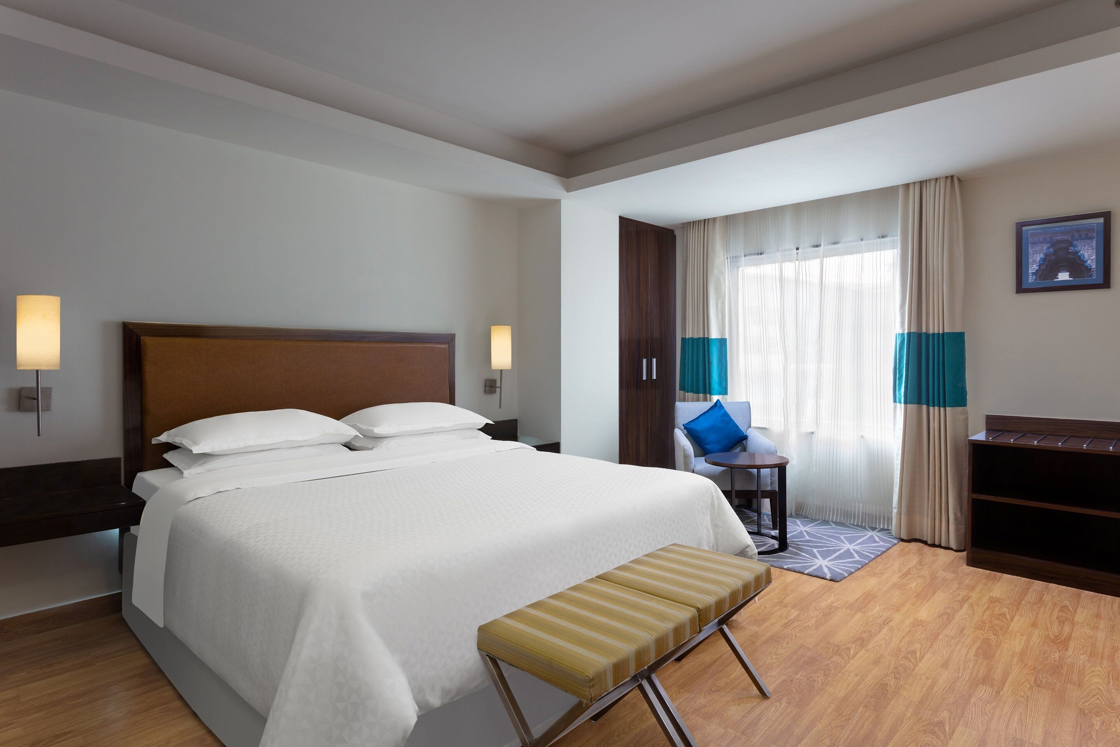 Une chambre d’hôtel avec un grand lit, des lampes de chevet, un banc, un bureau et une vue sur l’extérieur par une fenêtre munie de rideaux.