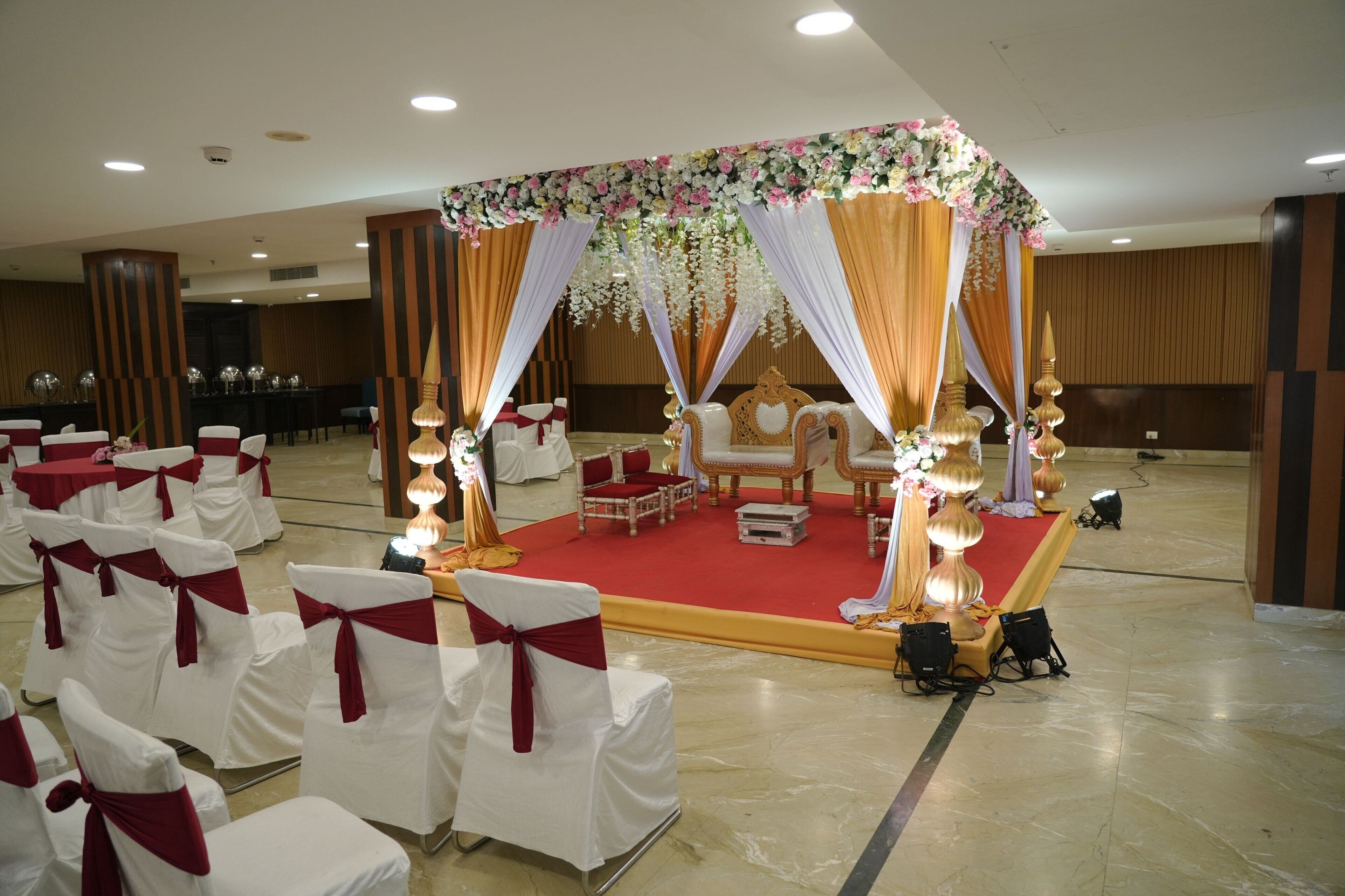 A banquet hall with rows of white chairs with red sashes, a stage with a red carpet, and a floral canopy.