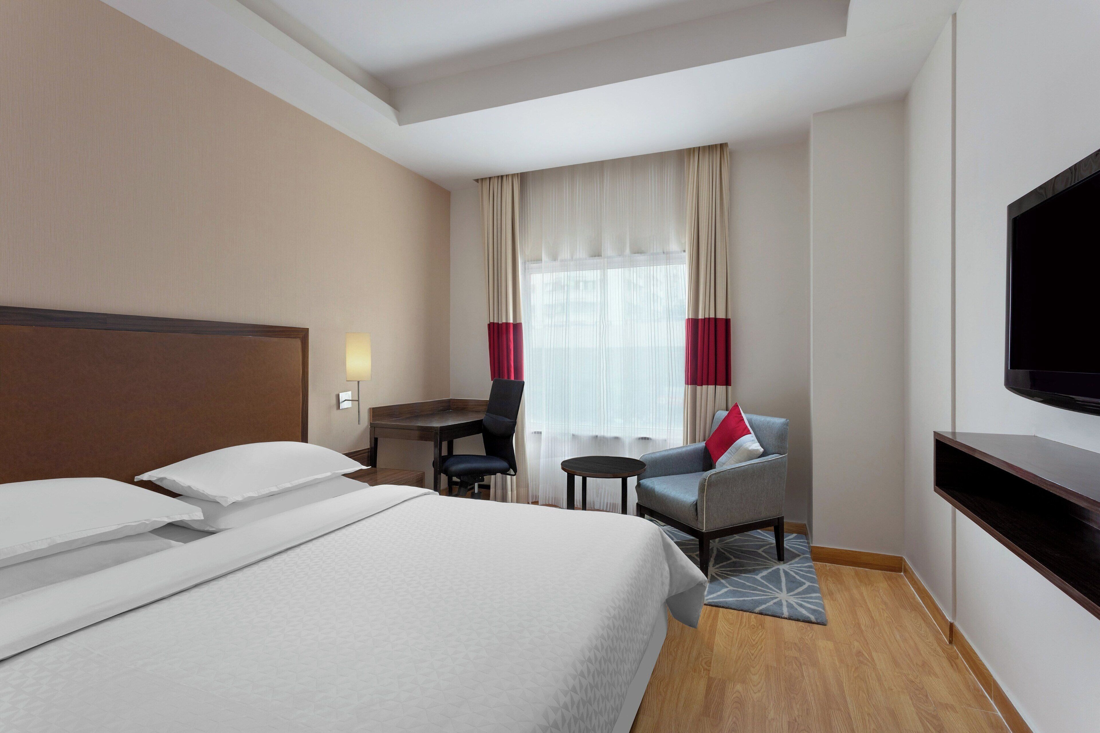 Habitació d'hotel amb un llit gran, un escriptori, una cadira, un televisor i una finestra amb cortines.