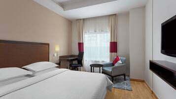 Habitació d'hotel amb un llit gran, un escriptori, una cadira, un televisor i una finestra amb cortines.