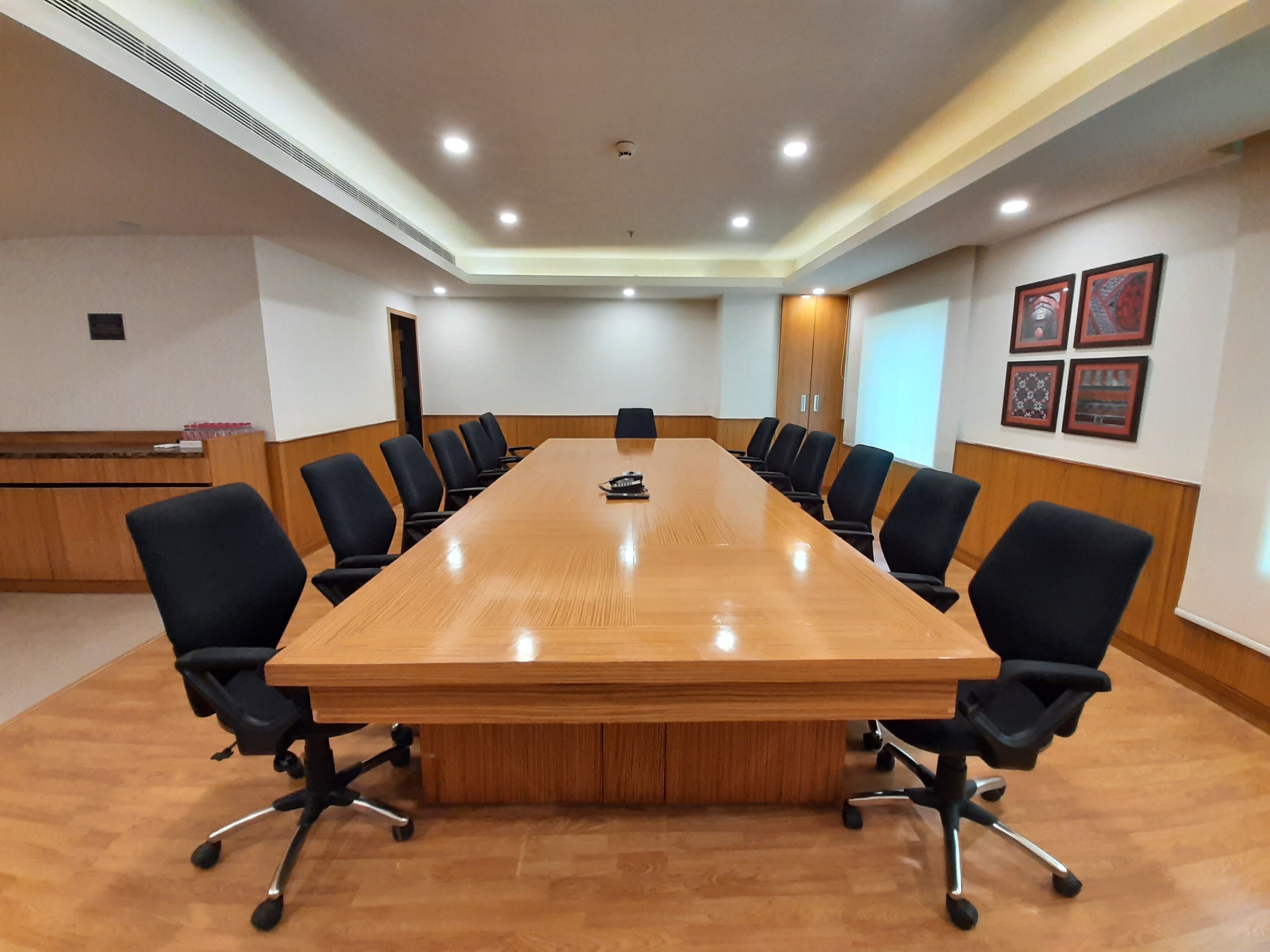A conference room with a long wooden table surrounded by black chairs, recessed ceiling lights, and framed pictures on the walls.