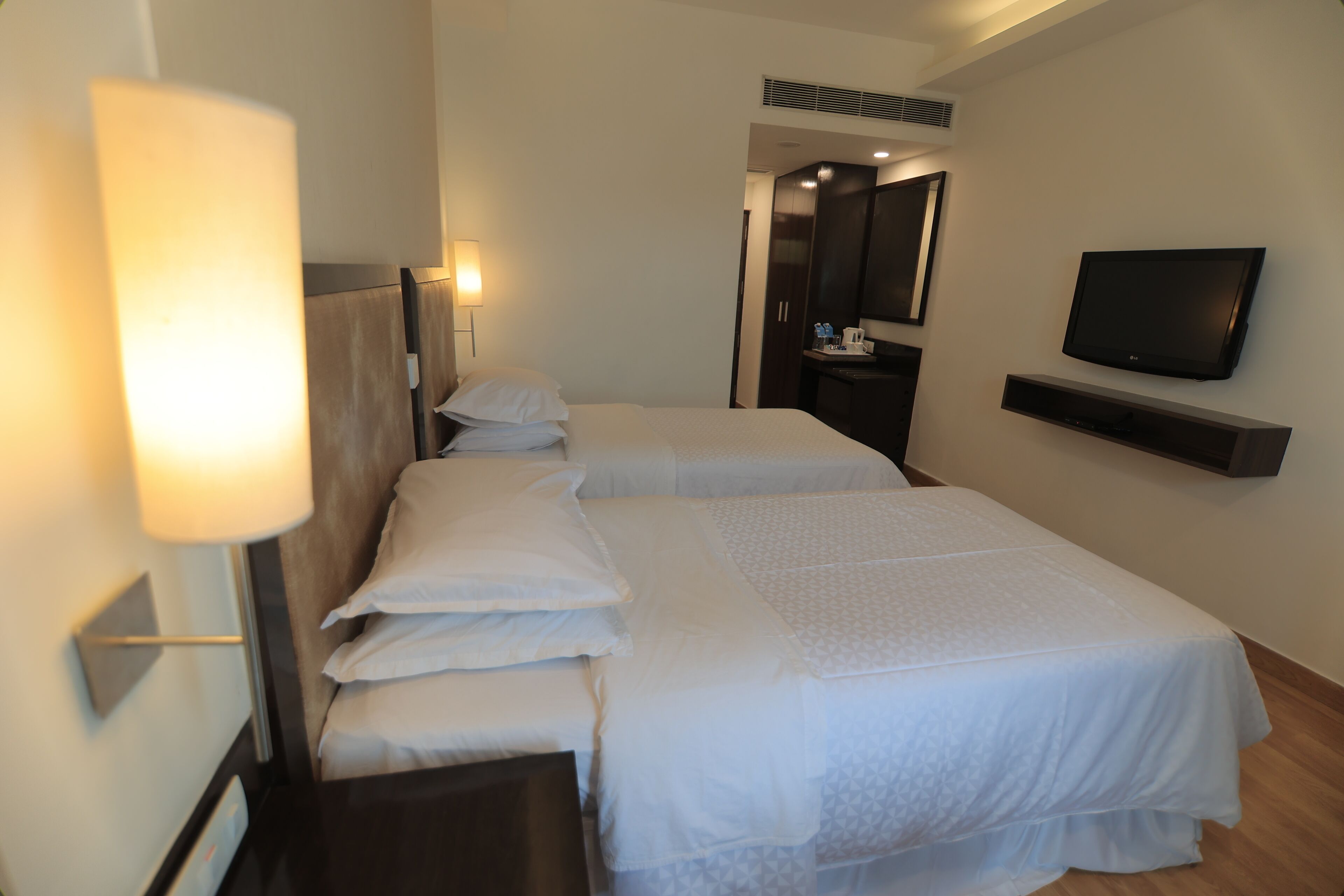 Habitación de hotel con dos camas, un televisor montado en la pared, un escritorio y una lámpara.