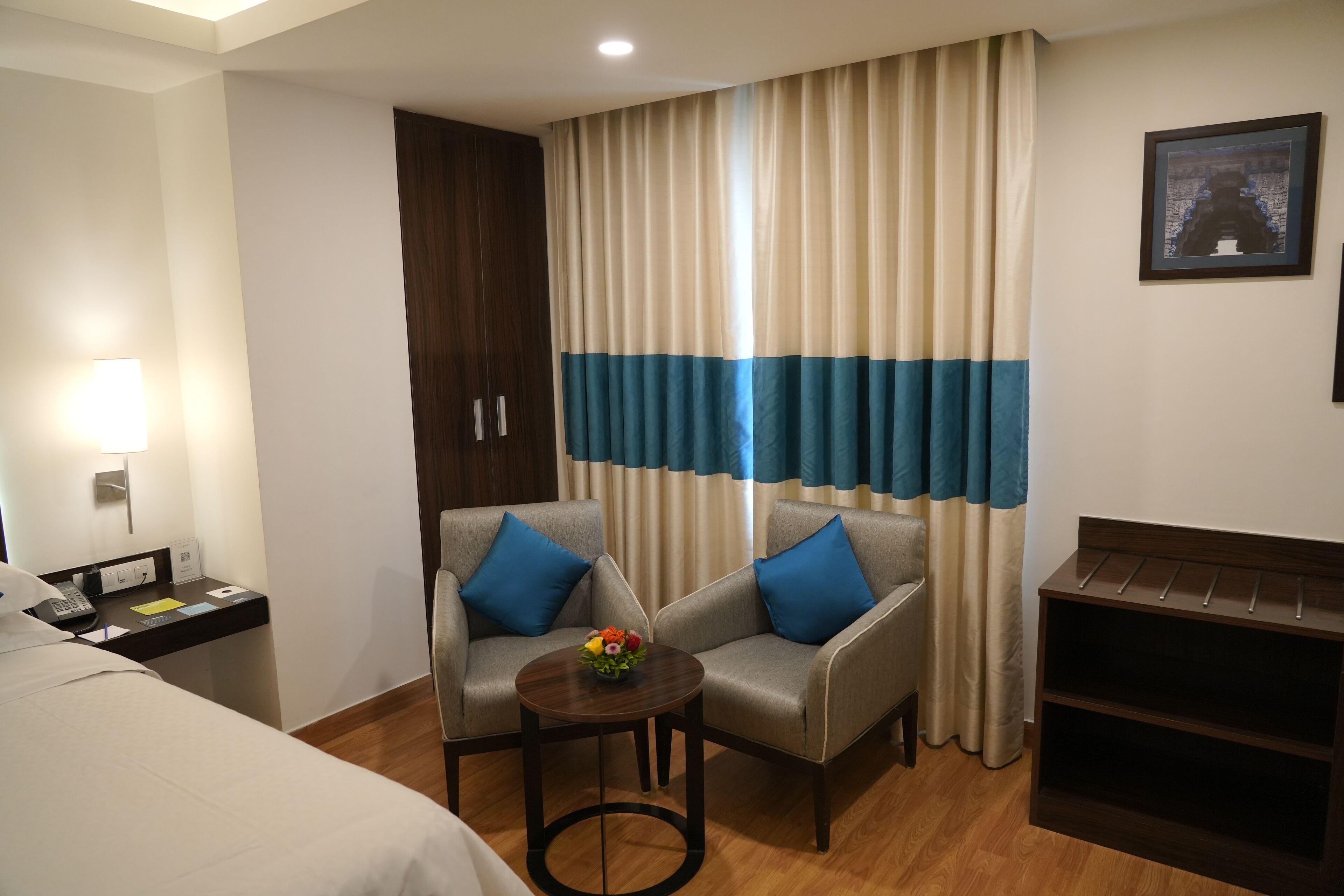 A hotel room with two armchairs, a small table with a flower vase, a bed, a nightstand with a phone, a lamp, a wall-mounted TV, and a large window with curtains.