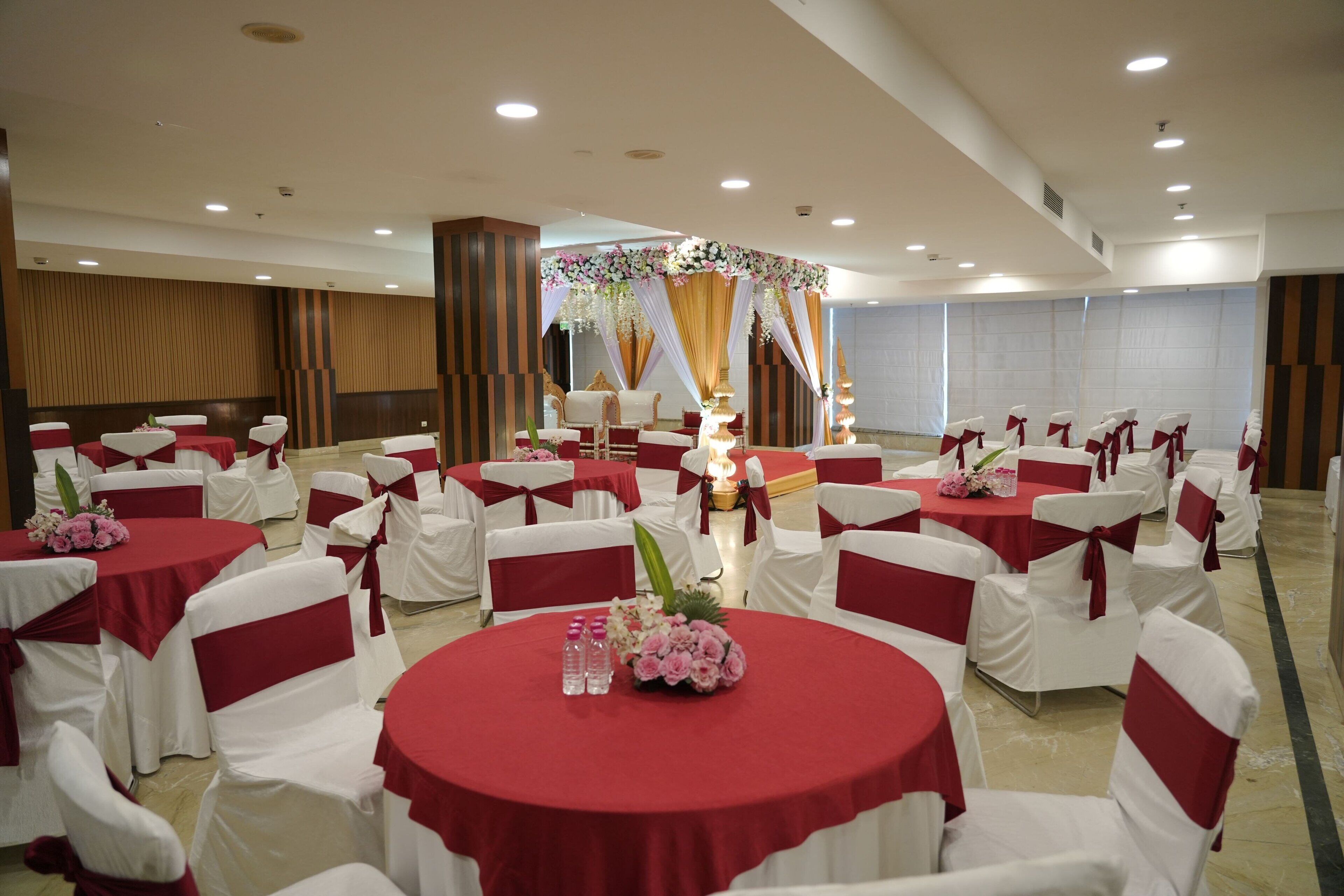 A banquet hall with round tables and white chairs, each with red sashes. Tables are adorned with floral centerpieces.