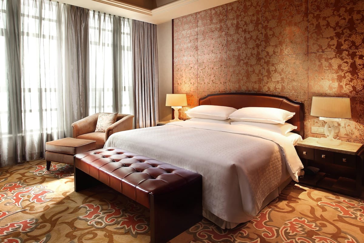 Une chambre d’hôtel avec un grand lit, un coin salon comprenant une chaise et un banc, et une moquette à motifs.