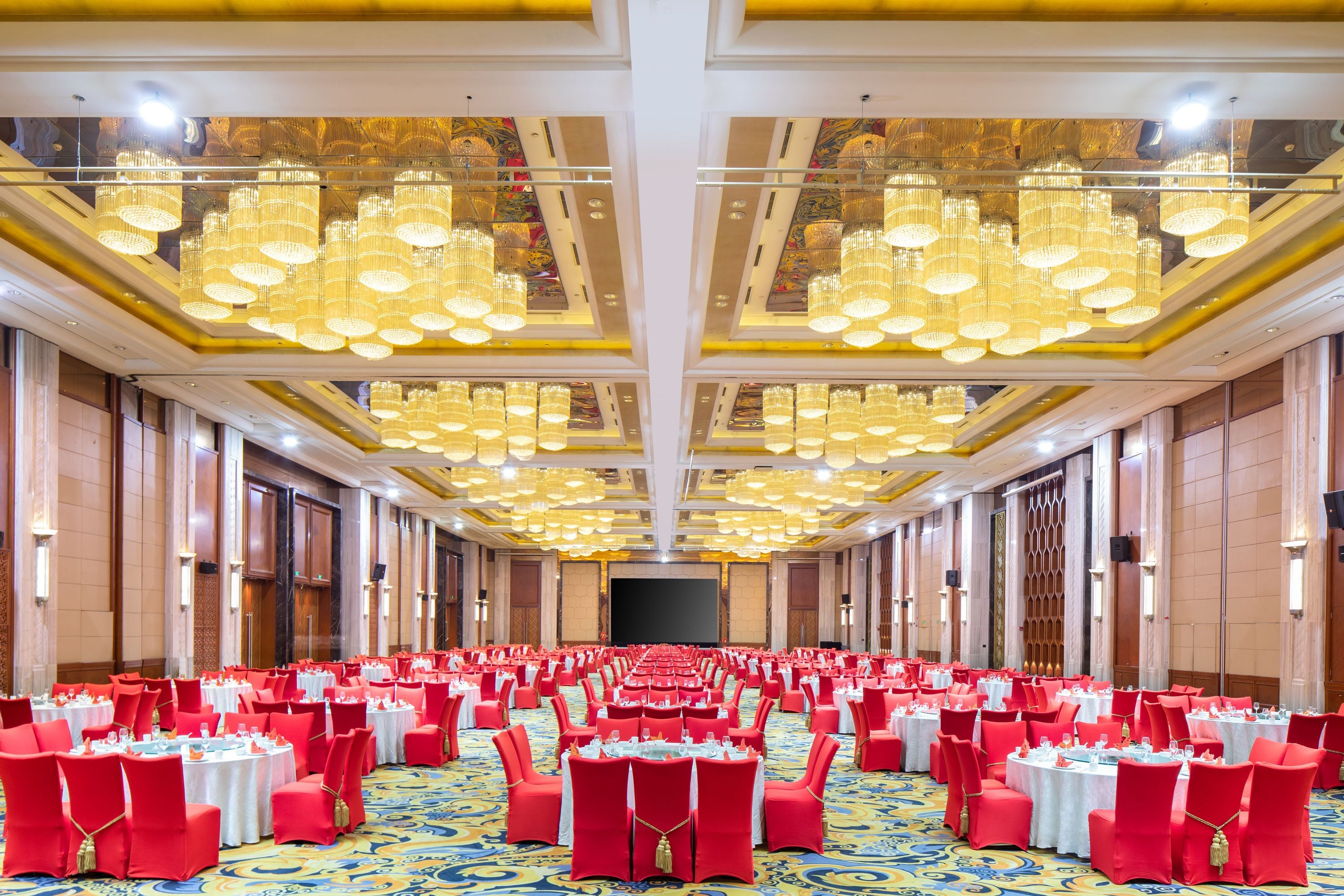 A banquet hall with rows of round tables, red chairs, and a large chandelier.