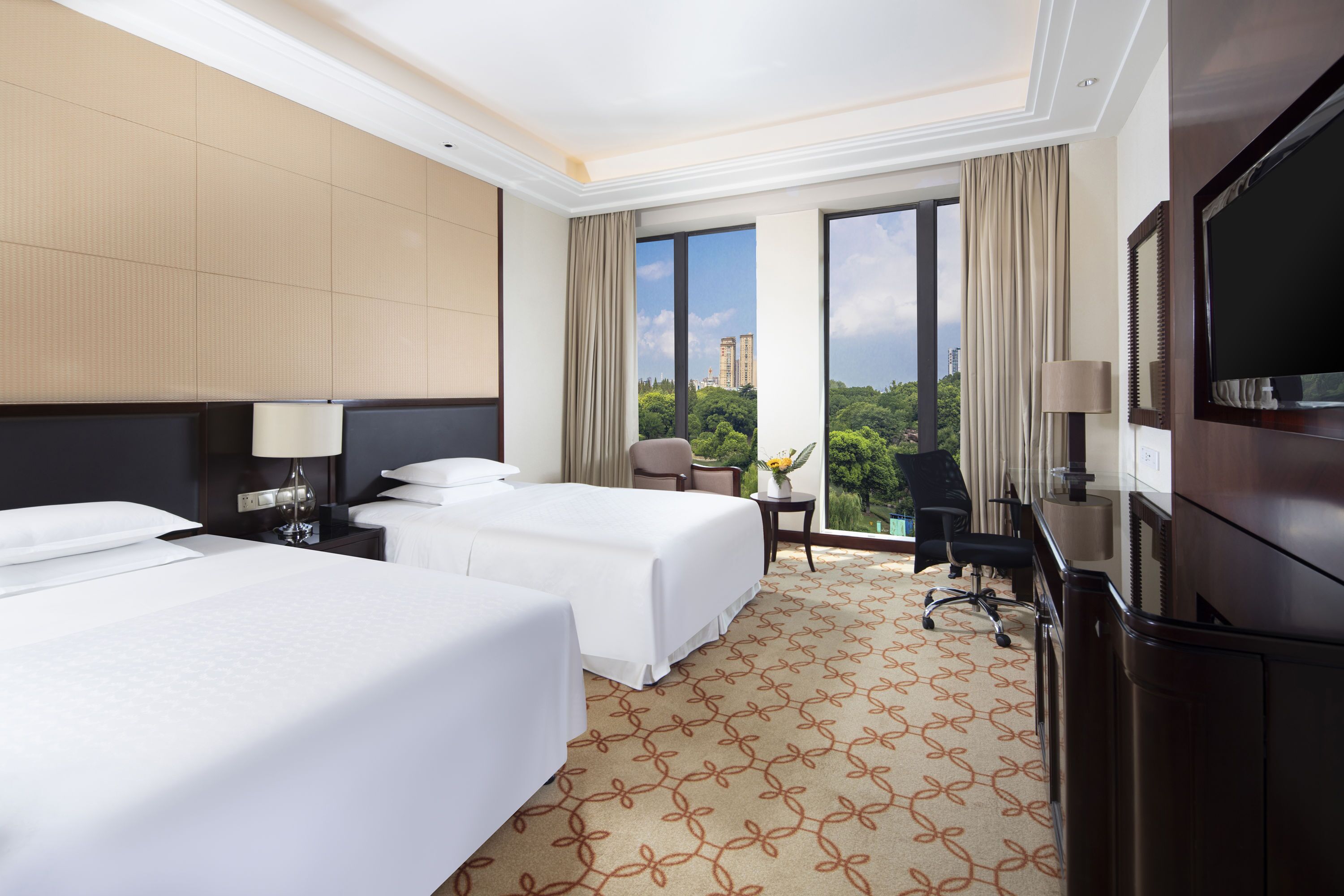 A hotel room with two beds, a desk, a TV, and a large window with a city view.