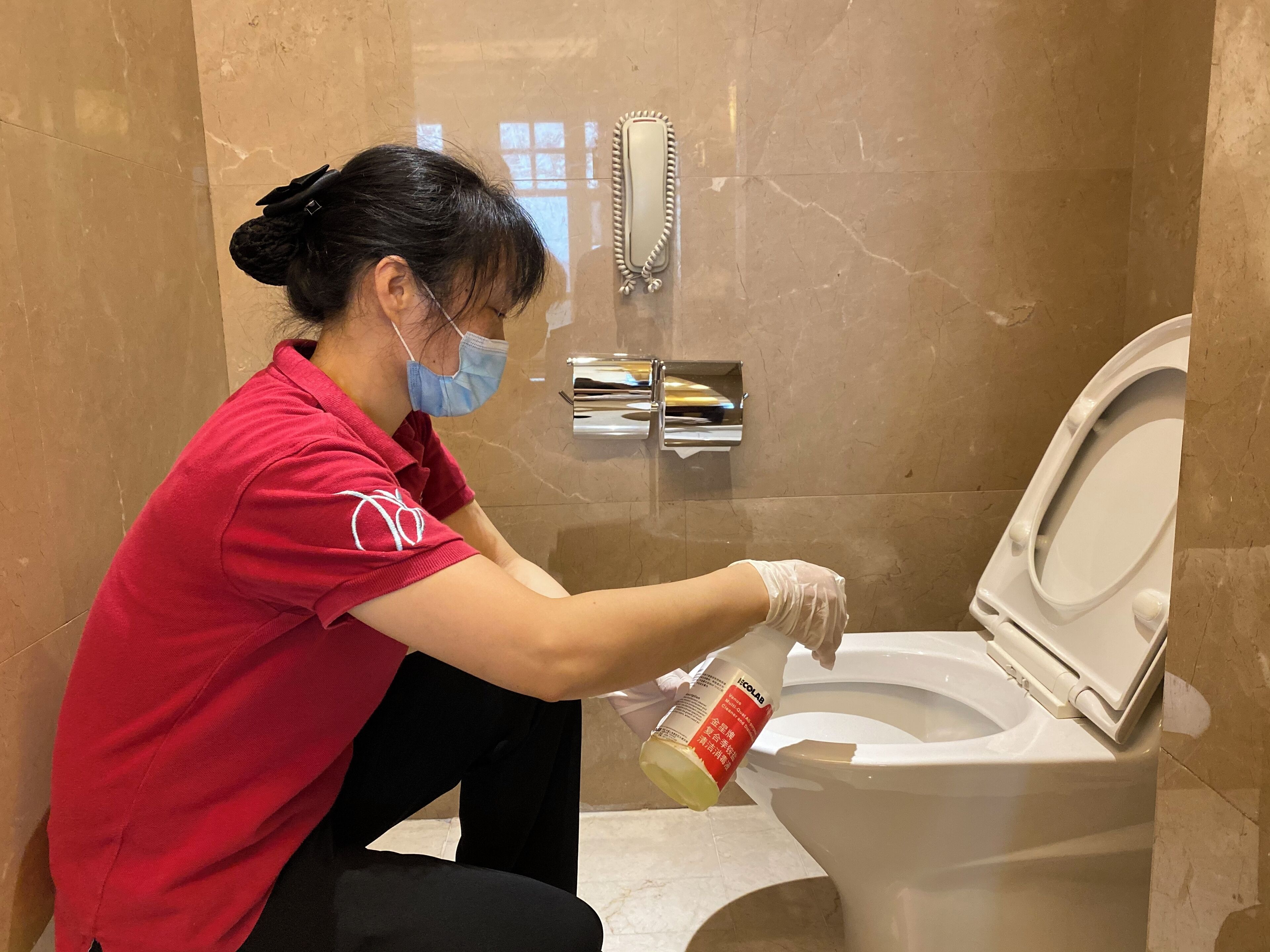 Una persona con uniforme rojo limpiando un inodoro en un baño con paredes de mármol y un espejo.