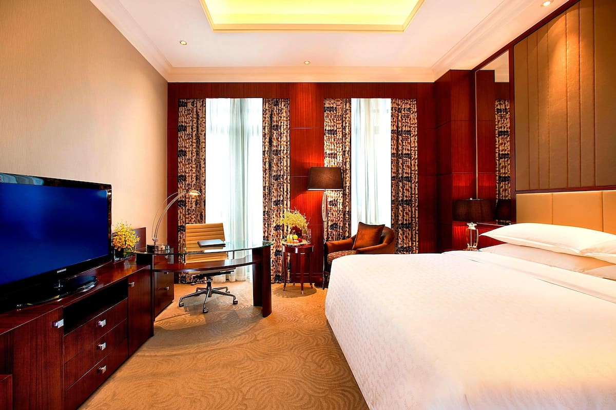 Une chambre d’hôtel dotée d’une grande télévision à écran plat, d’un bureau avec une chaise, d’un lit avec du linge de maison blanc et d’une grande fenêtre agrémentée de rideaux à motifs.