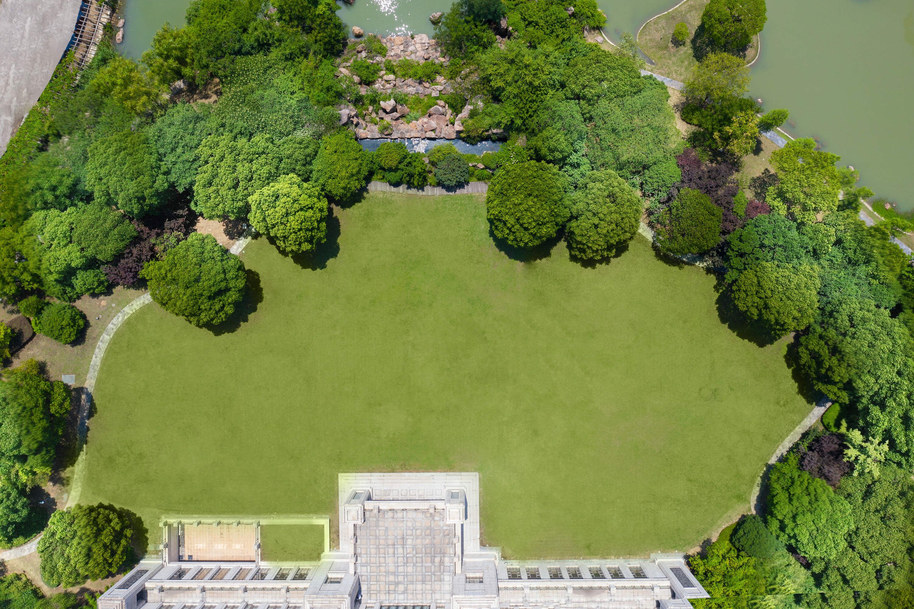 An aerial view of a park with a large open grassy area, surrounded by dense trees and a body of water.