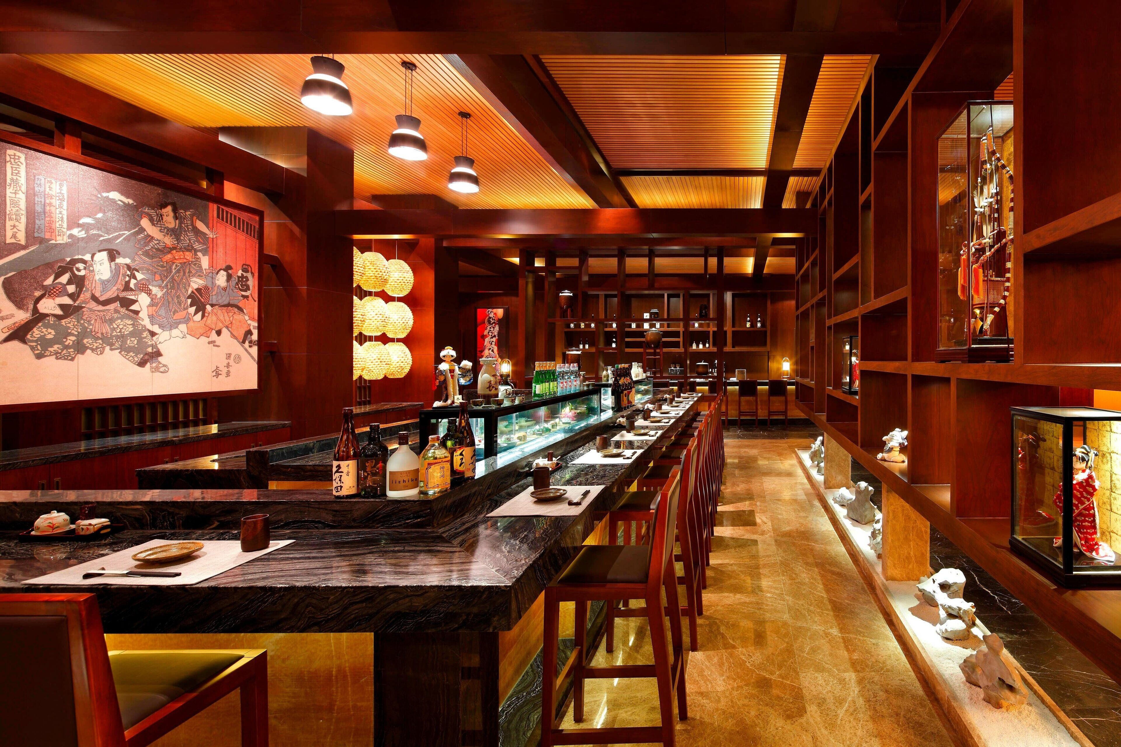 L’intérieur d’un restaurant japonais traditionnel, avec un long comptoir, des chaises en bois et une étagère où sont exposées des bouteilles de saké.