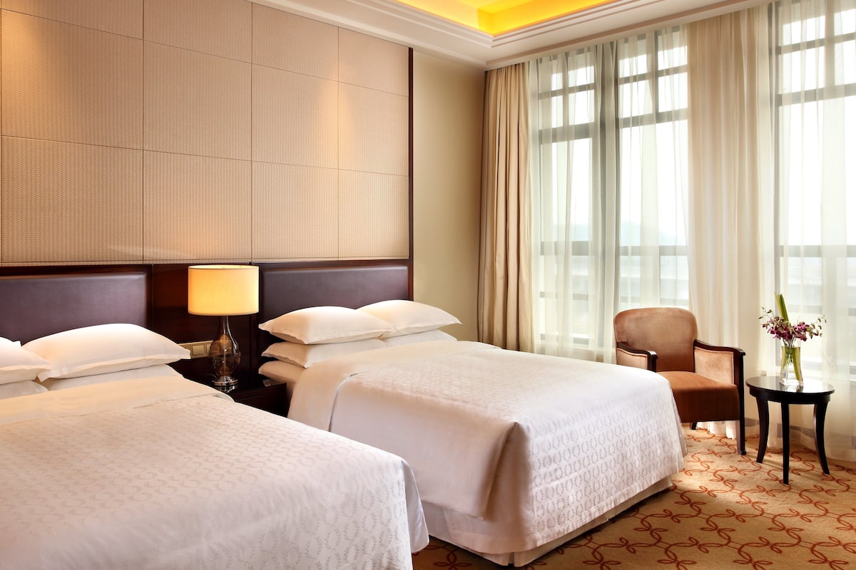 Une chambre d’hôtel avec deux lits, une table de chevet avec une lampe, une chaise, une petite table avec un vase et de grandes fenêtres avec des rideaux légers.