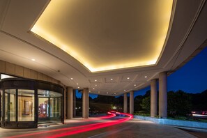Una entrada de hotel moderna con puerta giratoria de vidrio, un área techada amplia con columnas y vistas de la calle con estelas de luz.