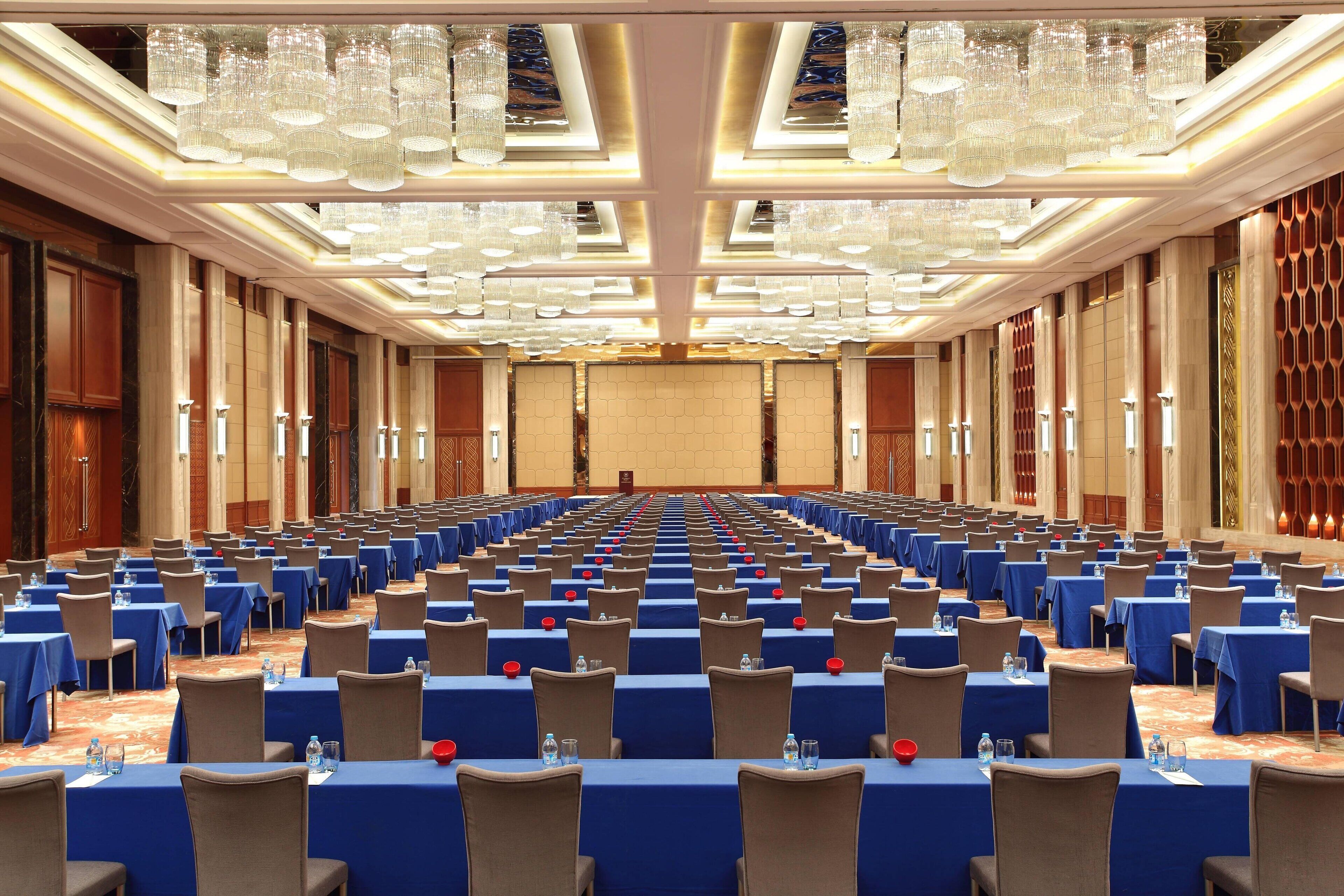 A large conference room with rows of tables and chairs, a stage at the front, and a high ceiling with chandeliers.
