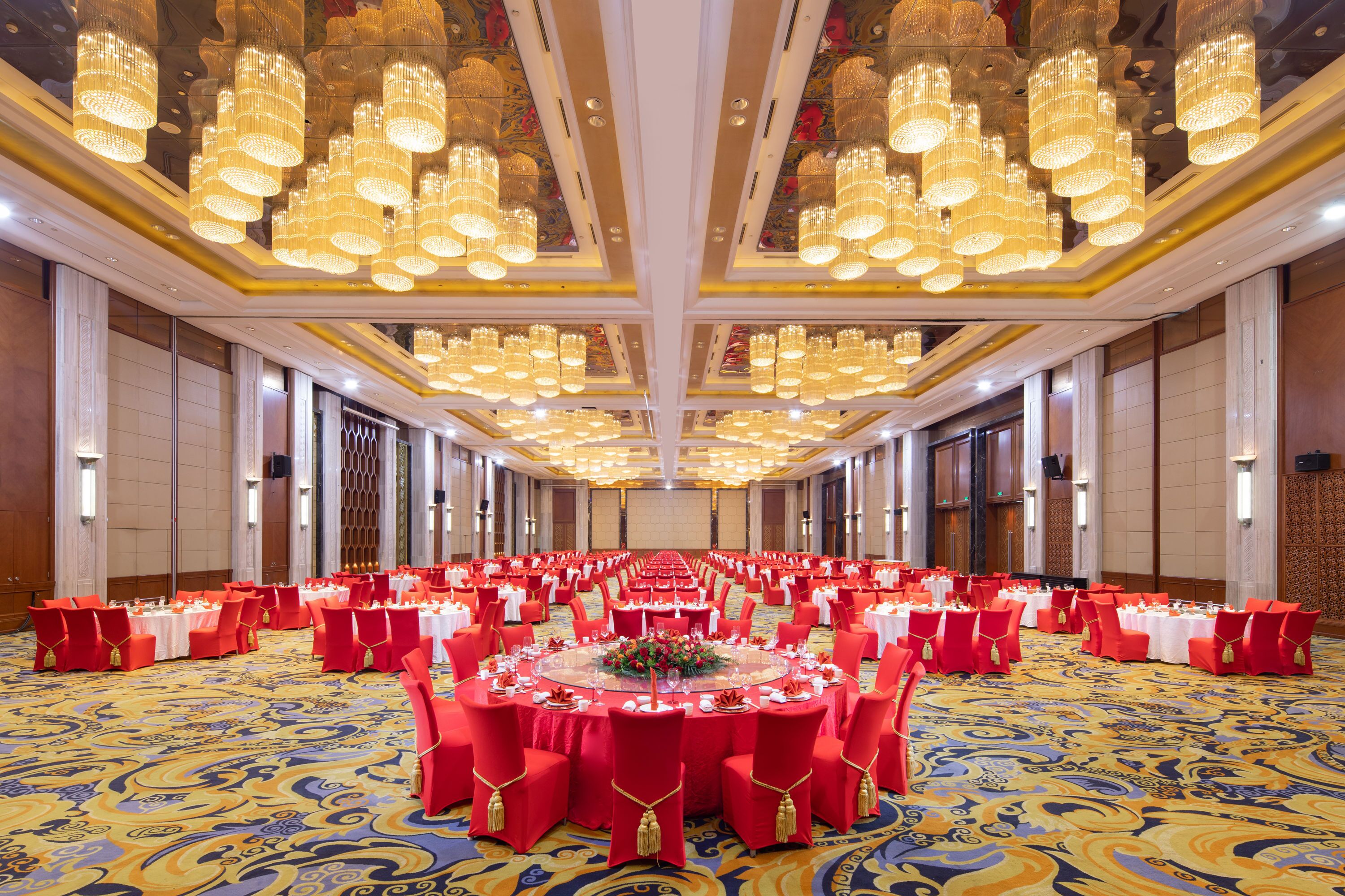 A banquet hall with round tables, red chairs, and chandeliers.