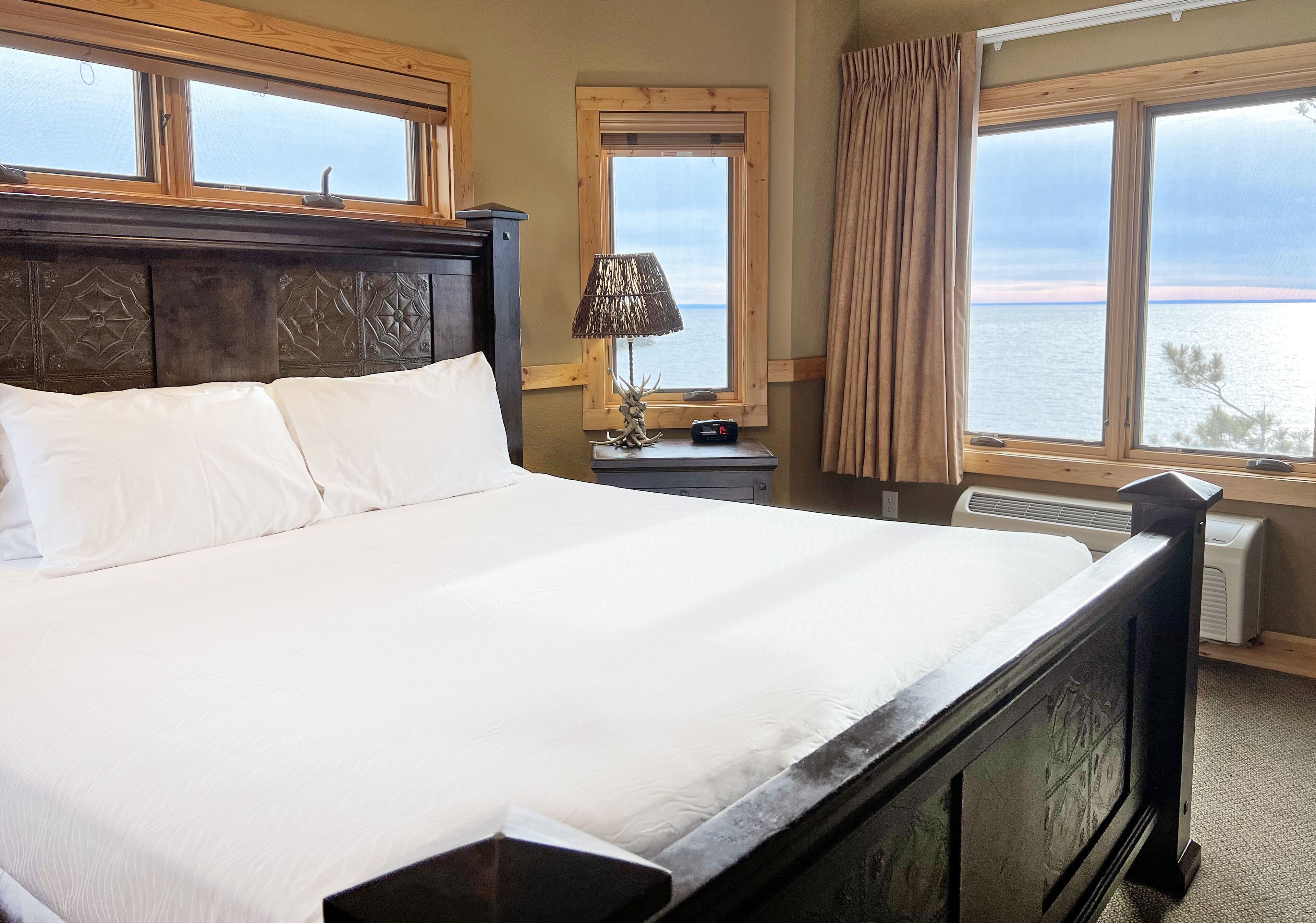 A hotel room with a large bed, a nightstand with a lamp, a window with a view of the sea, and a wooden headboard.