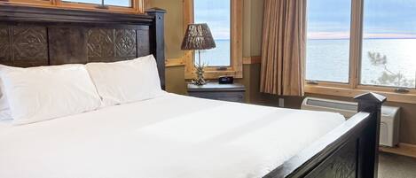 A hotel room with a large bed, a nightstand with a lamp, a window with a view of the sea, and a wooden headboard.