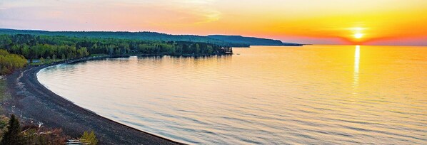 Un paysage côtier avec une ligne de rivage incurvée, une zone boisée et un coucher de soleil sur l’eau.