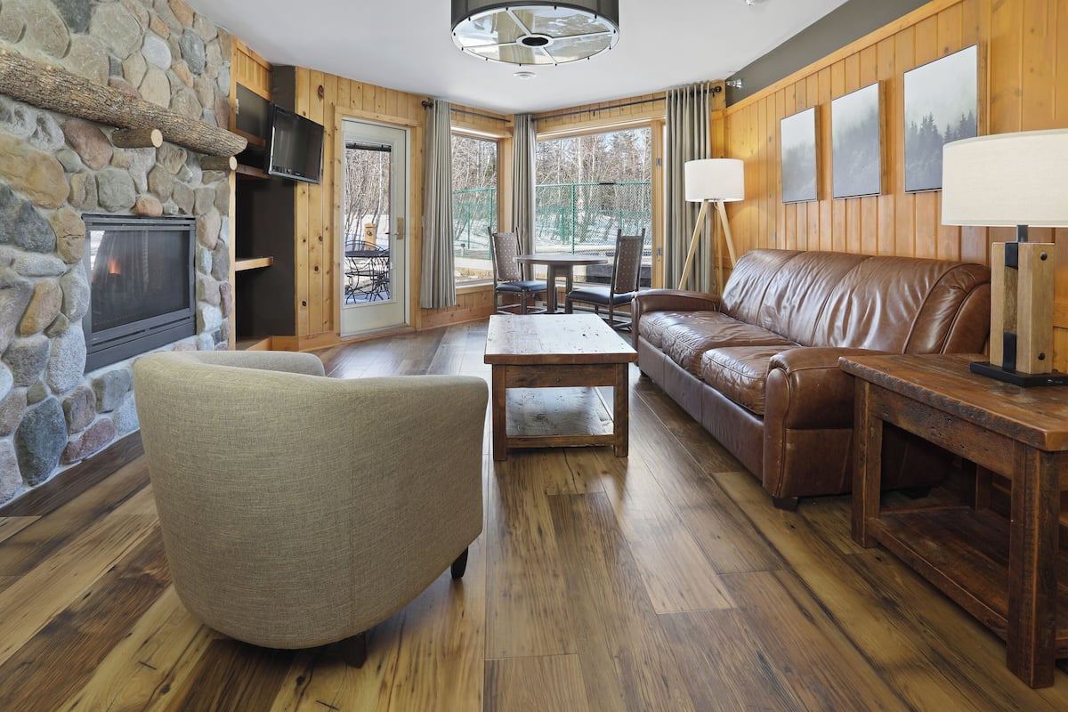 A cozy living room with a stone fireplace, leather sofa, wooden flooring, and large windows.