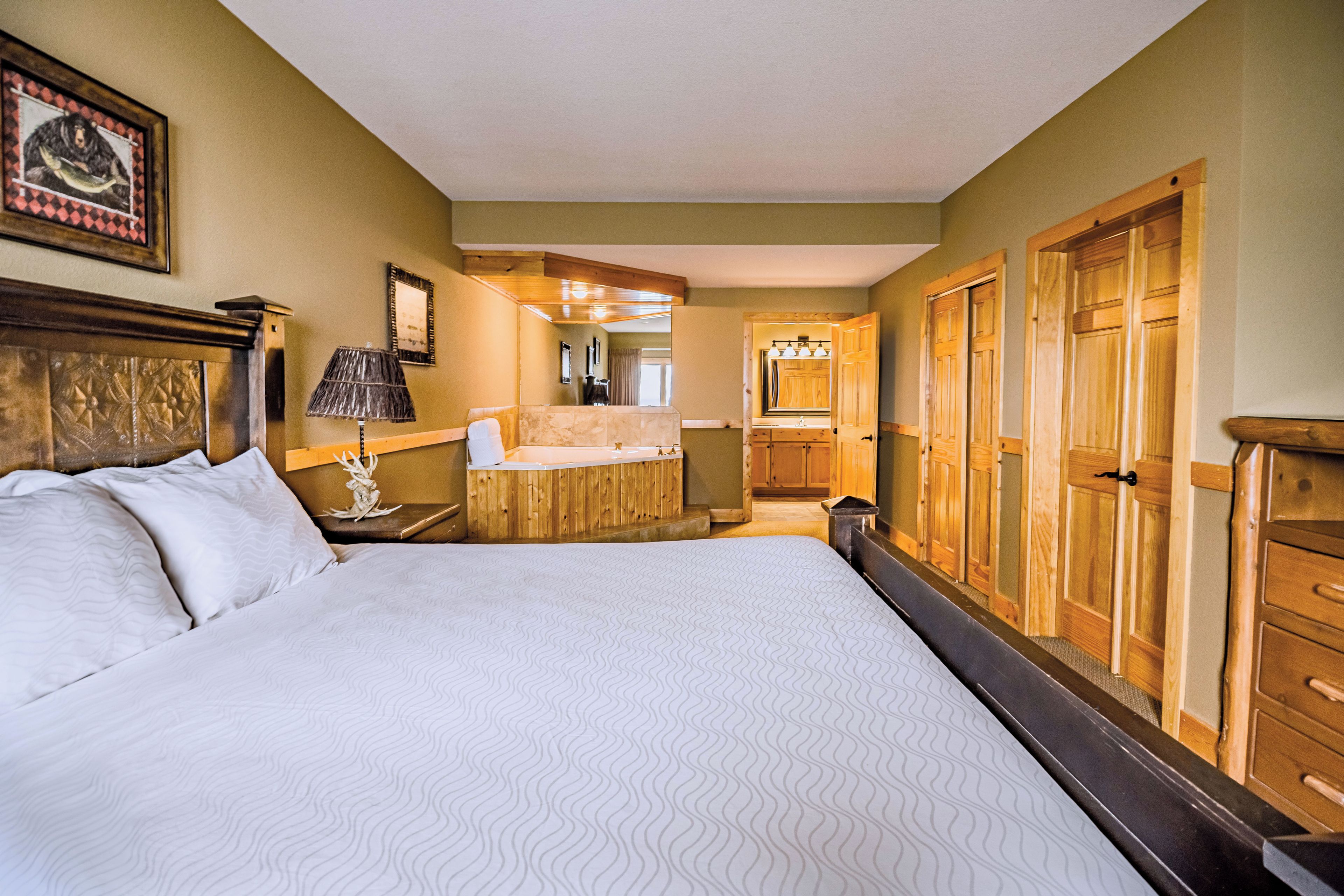 A hotel room with a large bed, wooden wardrobe, a small bathroom with a mirror, and a wooden cabinet.