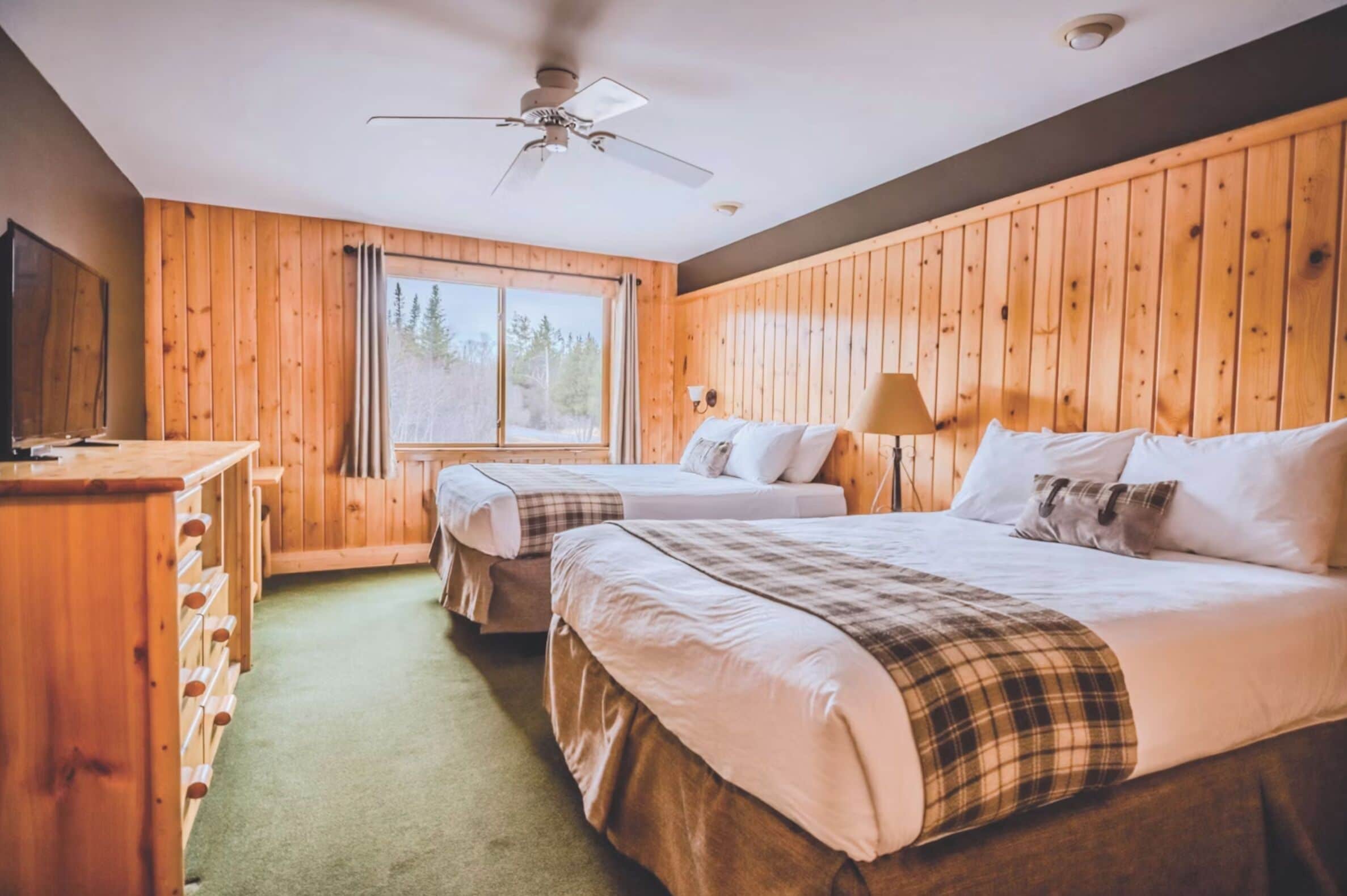 Une chambre avec deux lits, un ventilateur de plafond, une fenêtre avec des rideaux et un mur en bois.