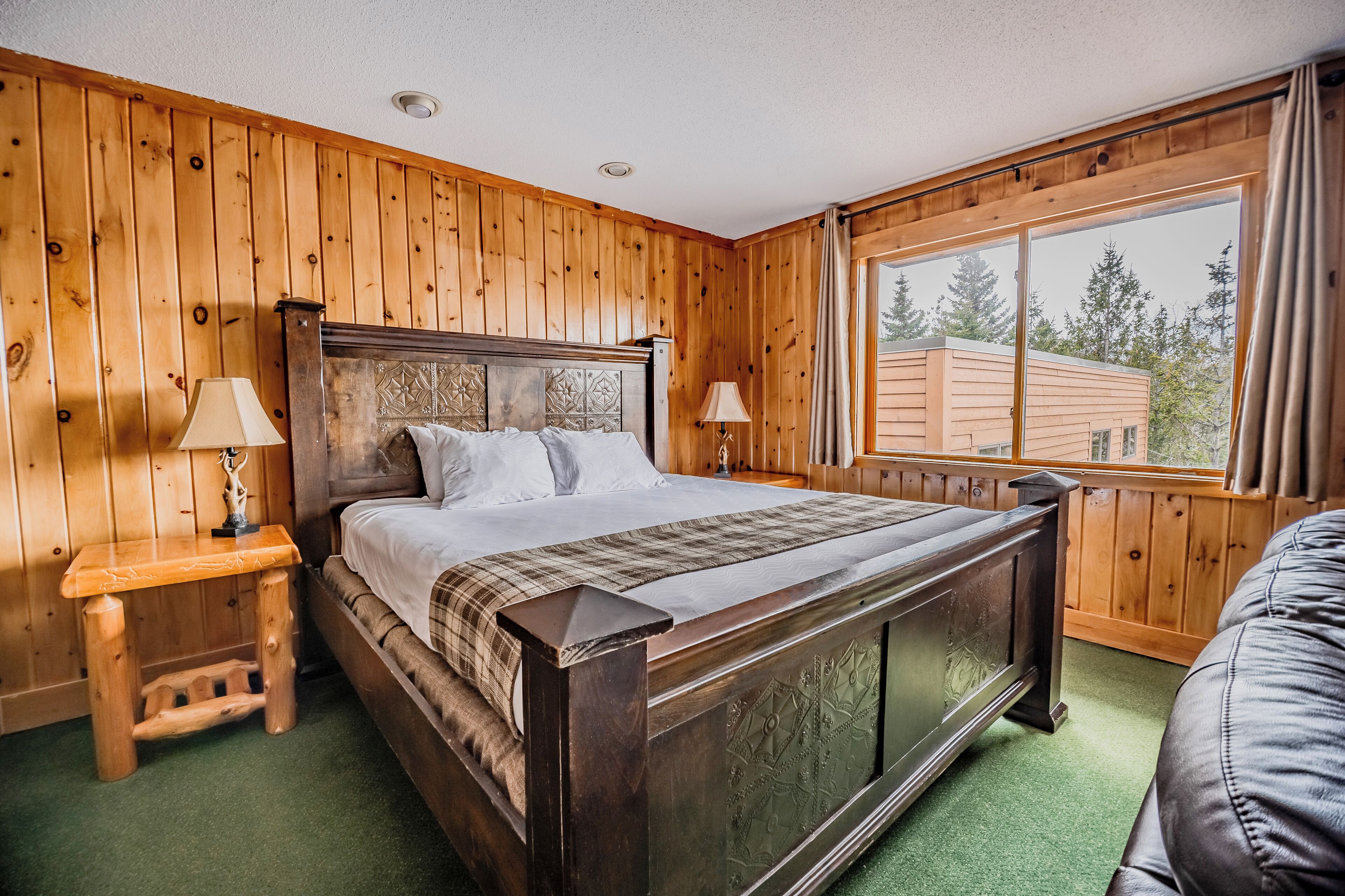 Un dormitorio con paneles de madera, una cama grande, dos mesitas de noche con lámparas y una ventana con vistas a árboles.