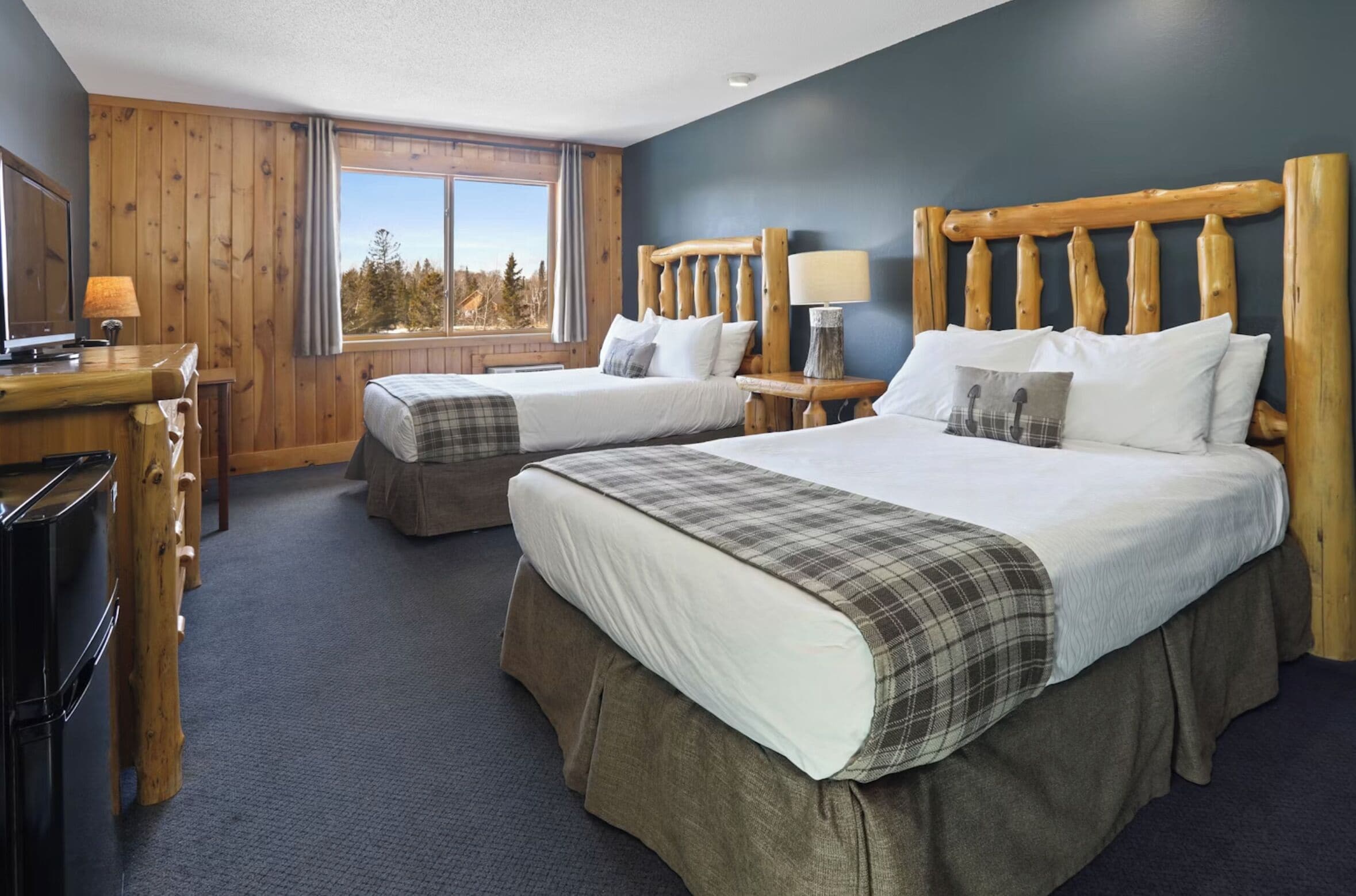 A hotel room with two beds, wooden headboards, a desk with a lamp, and a window with a view of trees.